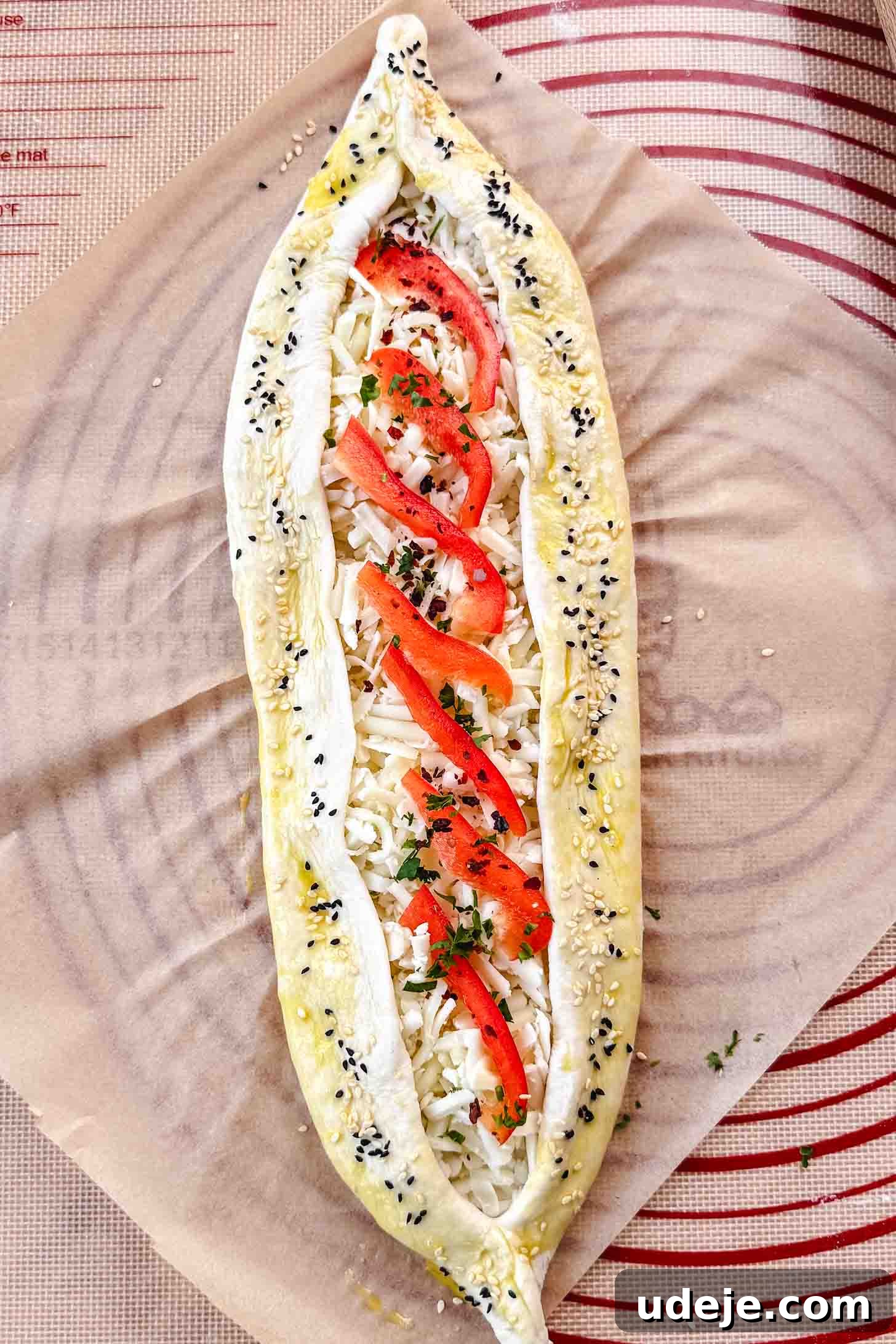 Customizable Homemade Turkish Pide 13 A boat-shaped dough filled with shredded white cheese, adorned with chopped parsley and vibrant slices of red pepper, along with nigella and sesame seeds, prepared on parchment paper.