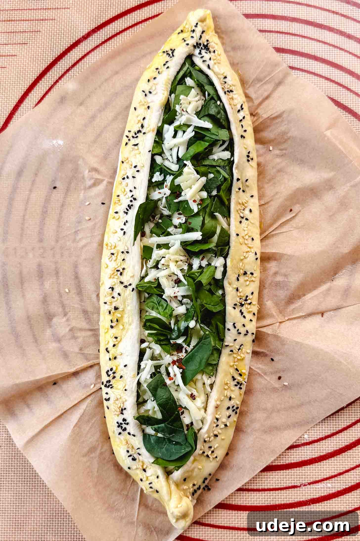 Customizable Homemade Turkish Pide 14 A boat-shaped dough filled with a delicious combination of shredded white cheese and finely chopped spinach, elegantly sprinkled with nigella and sesame seeds, laid out on parchment paper.