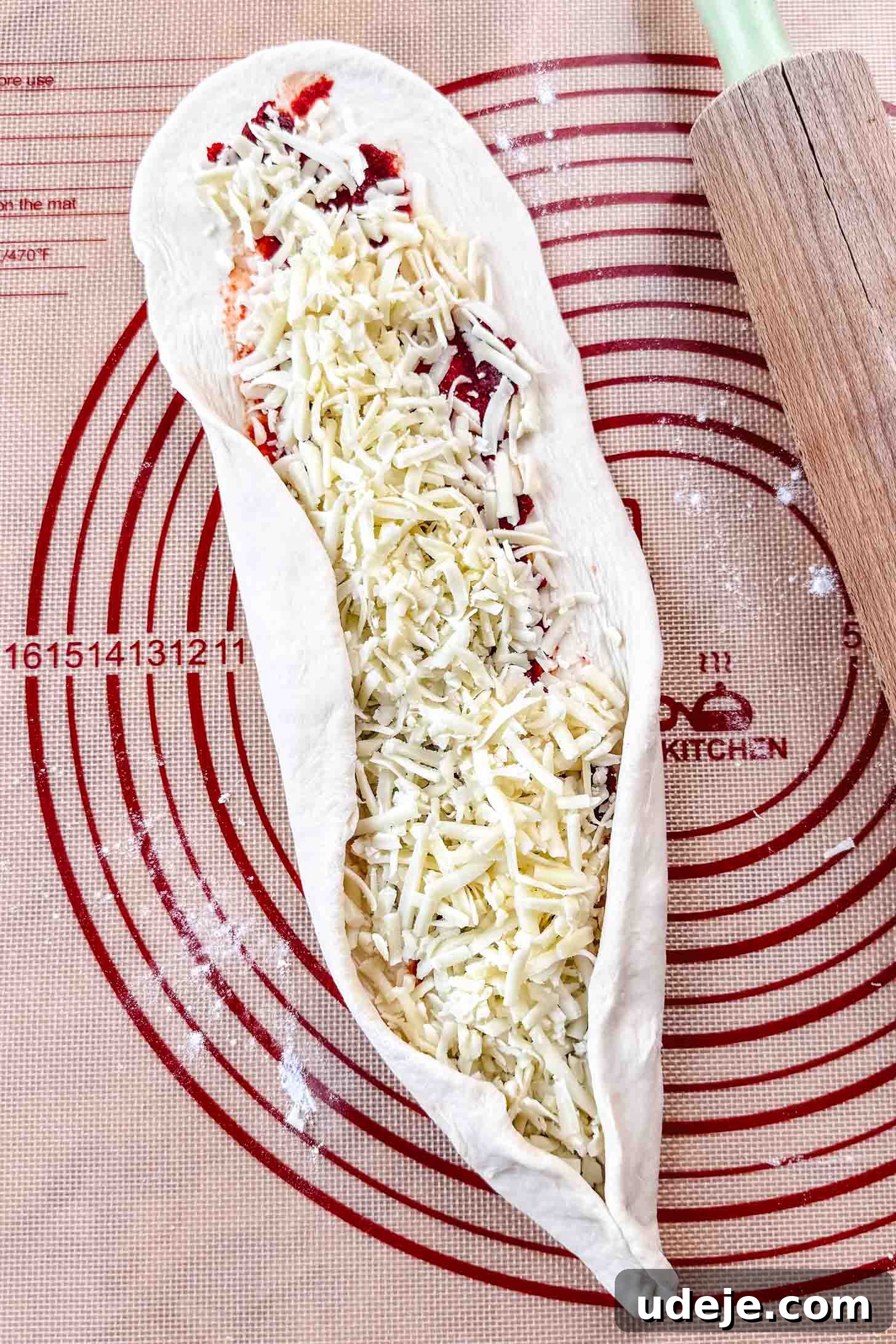 Customizable Homemade Turkish Pide 10 An oval-shaped dough, spread with red pepper paste and topped with shredded cheese, is being skillfully folded up at the bottom edge on a red-circled Silpat mat, with a wooden rolling pin nearby.