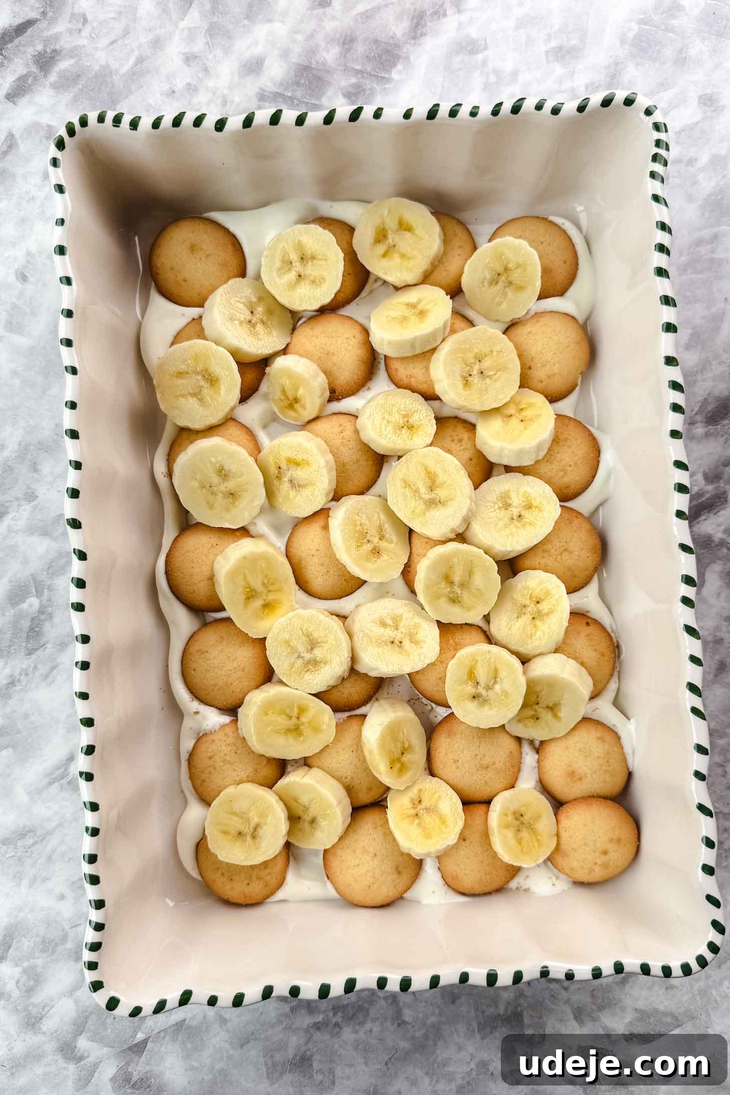 Magnolia Bakery's Irresistible Banana Pudding 11 White baking dish with green stripes on the edge with cream filling spread at the bottom, circular cookies and bananas on a marble background.