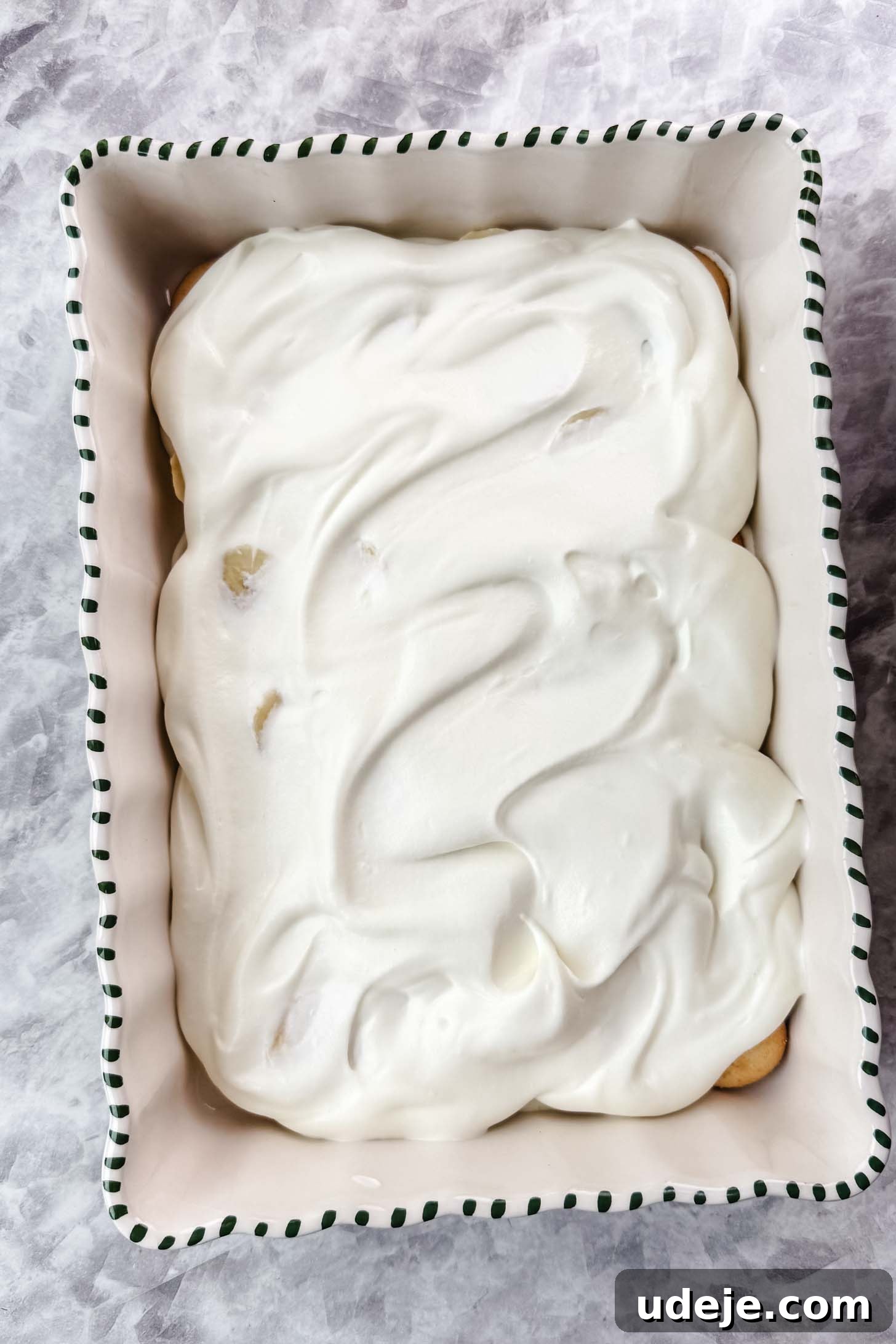 Magnolia Bakery's Irresistible Banana Pudding 12 White baking dish with green stripes on the edge with cream filling spread on top on a marble background.