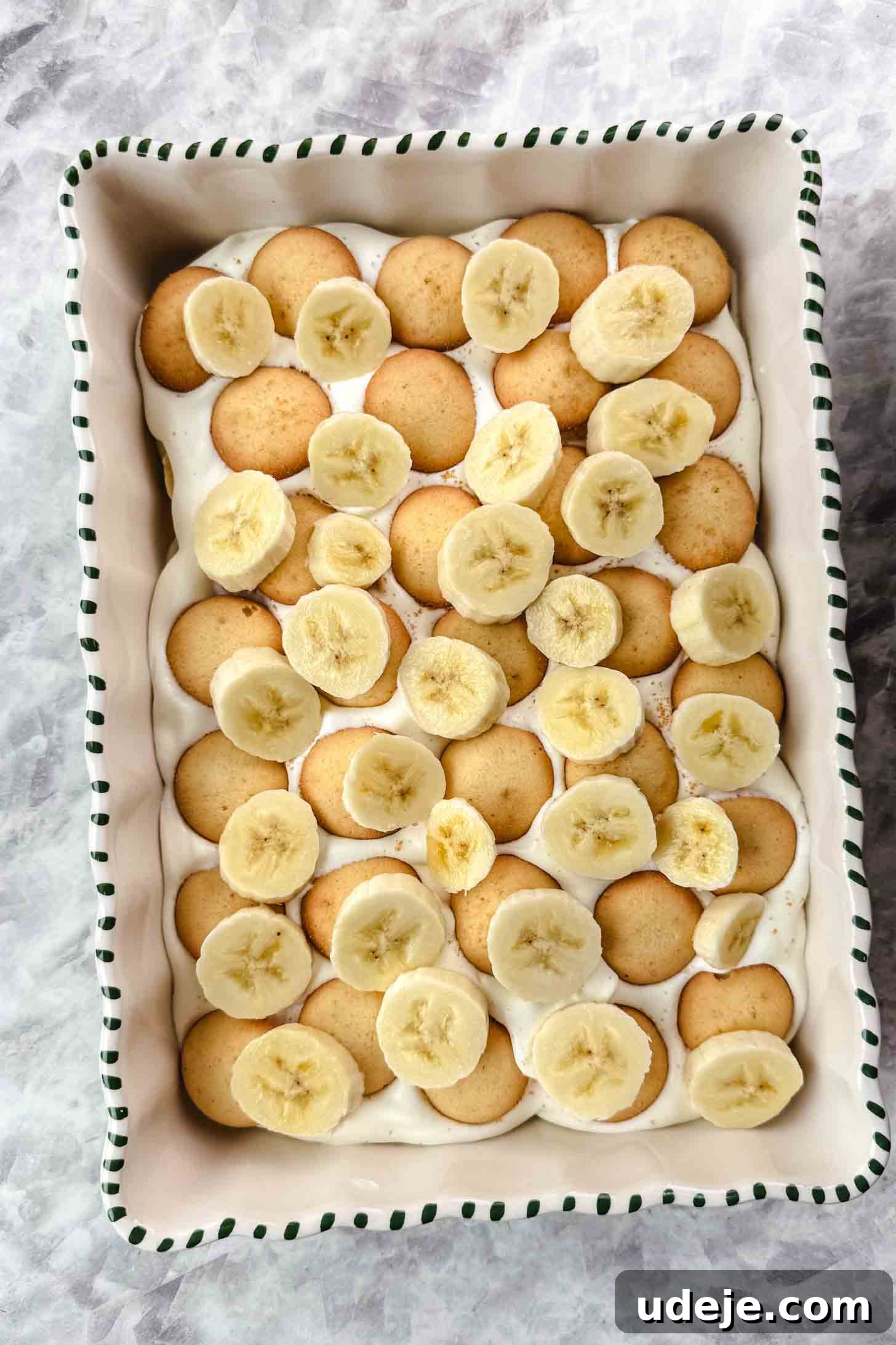 Magnolia Bakery's Irresistible Banana Pudding 13 White baking dish with green stripes on the edge with cream filling spread on top of layers of circular cookies and sliced bananas on a marble background.