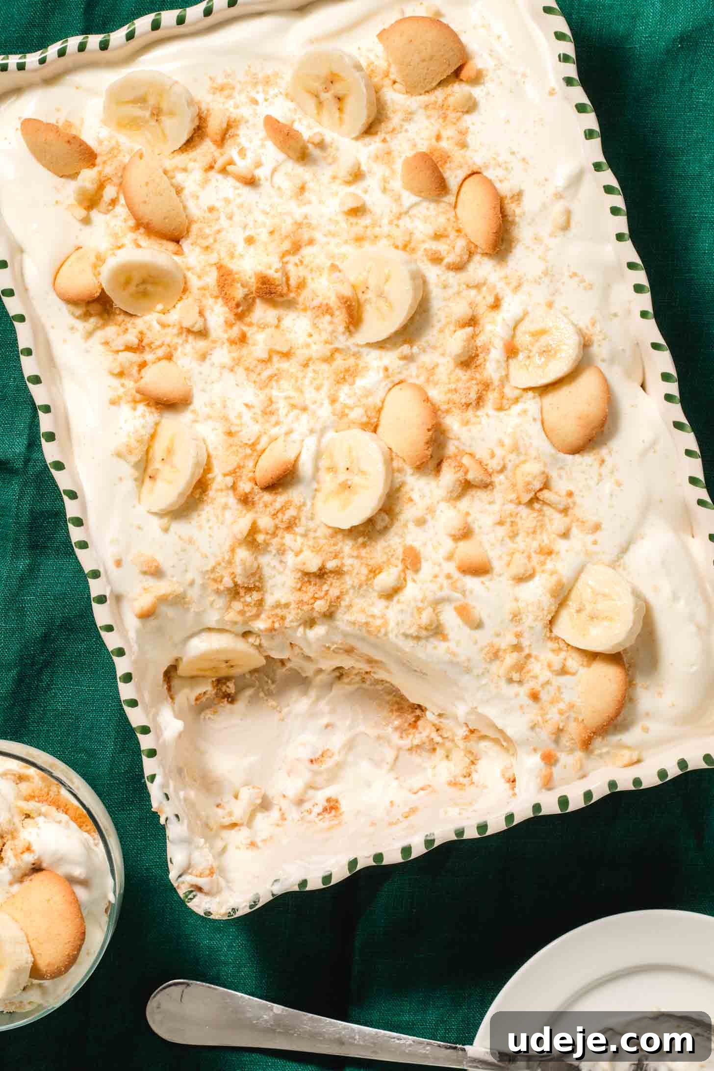 Magnolia Bakery's Irresistible Banana Pudding 14 Creamy layered dessert with cream, sliced bananas and circular cookies in a white baking dish with green stripes on the edge. A portion has been dished out into a glass bowl beside it with a white plate and a serving spoon at the side on a dark green tablecloth.