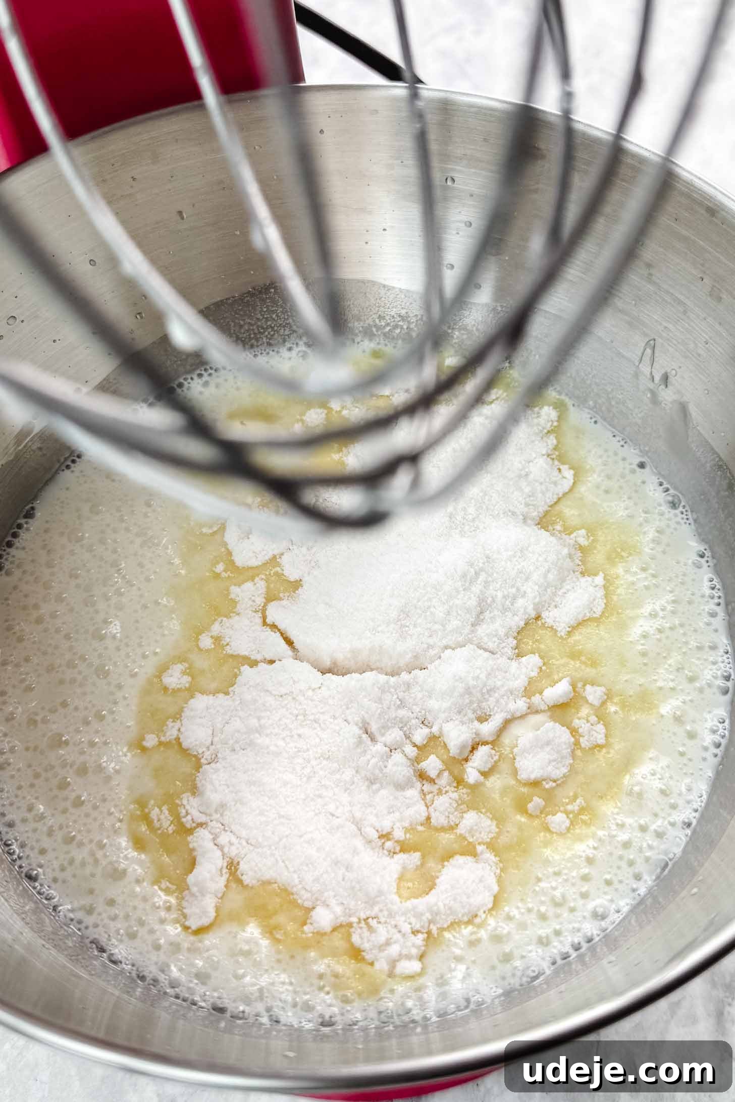 Magnolia Bakery's Irresistible Banana Pudding 4 Silver bowl of a stand mixer with water, vanilla pudding powder and condensed milk with the whisk attachment above on a marble background.