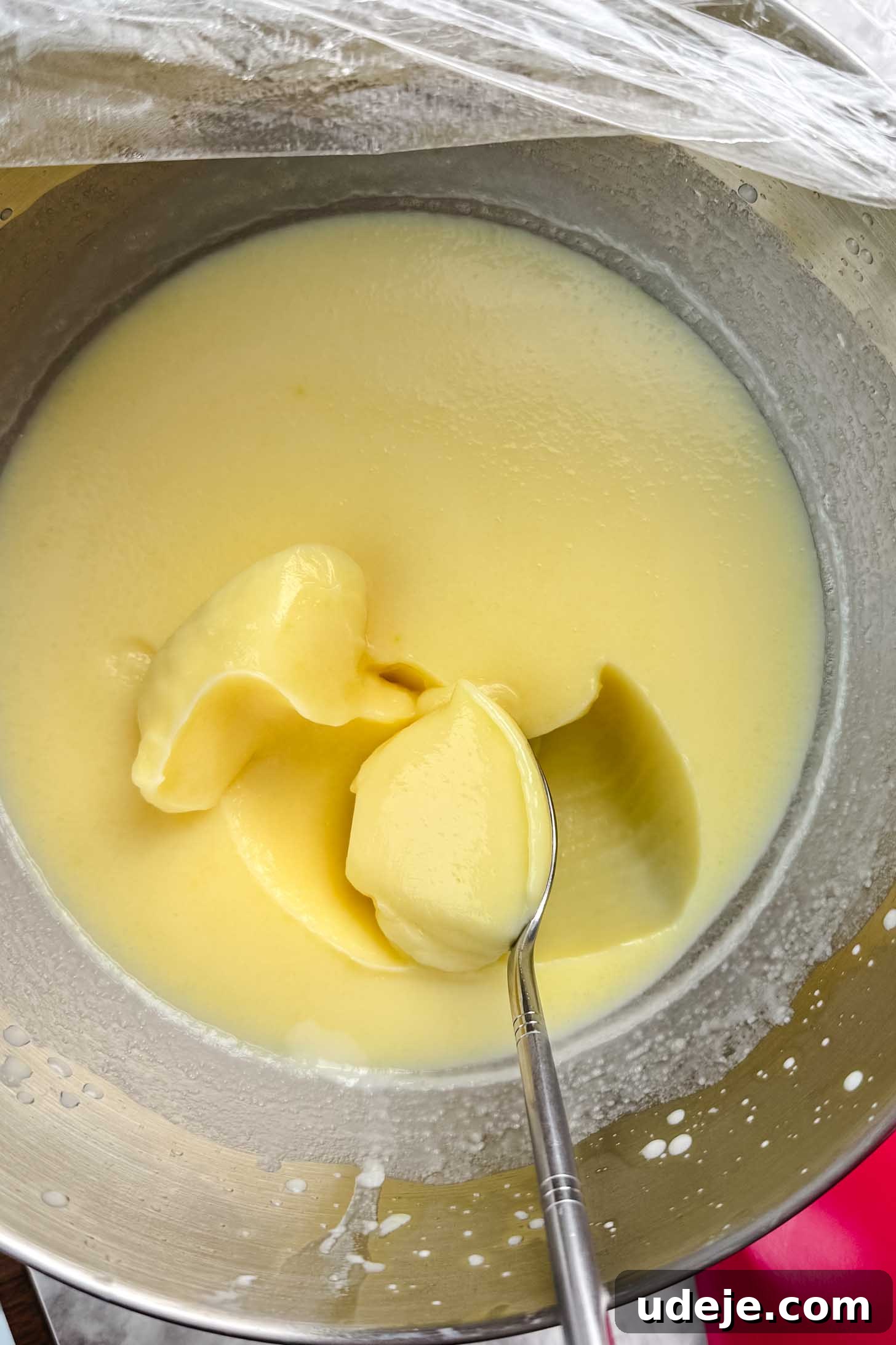 Magnolia Bakery's Irresistible Banana Pudding 6 A yellow pudding in the silver bowl of a stand mixer with a spoon in it and plastic wrap at the top corner.