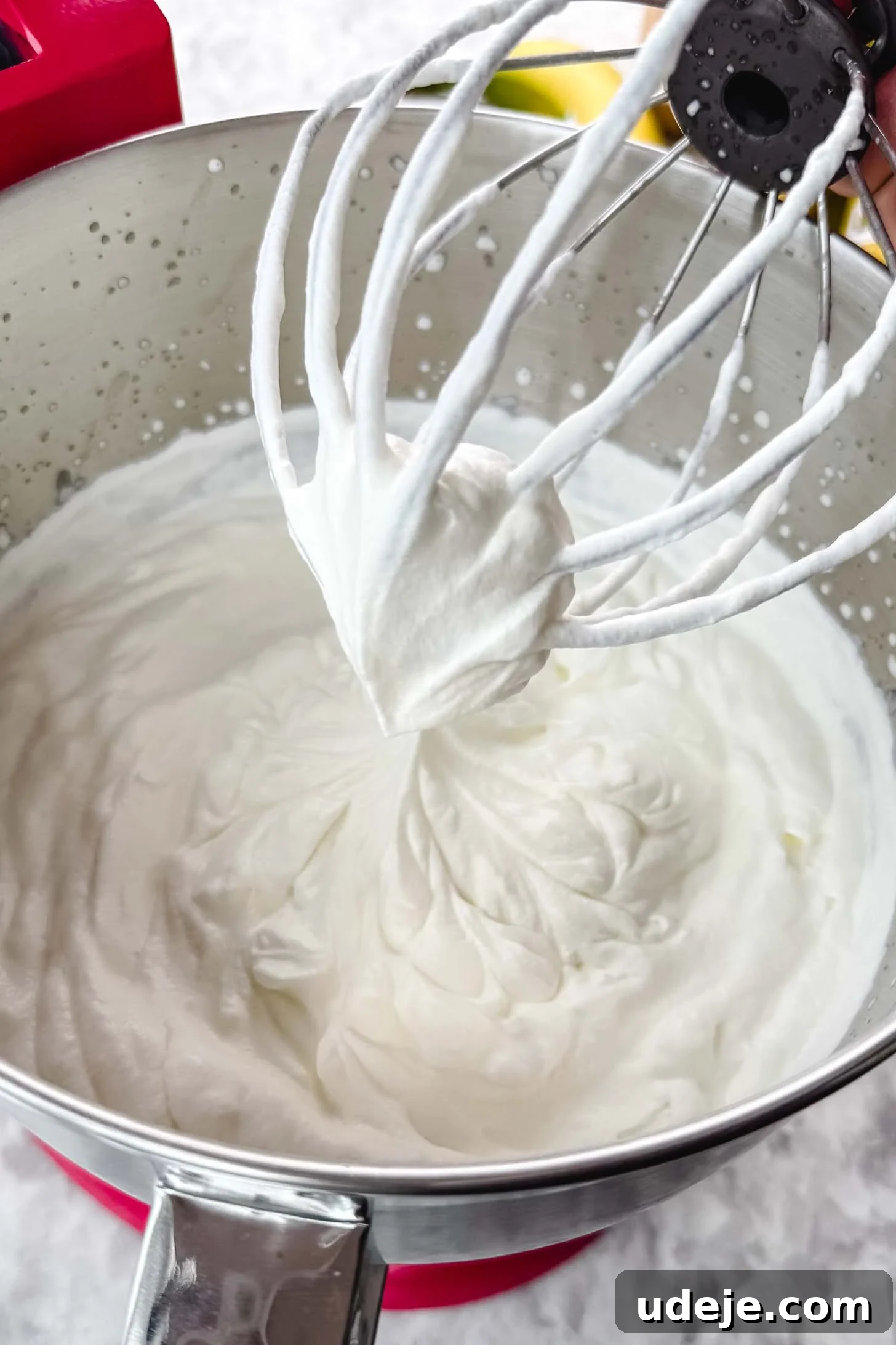 Magnolia Bakery's Irresistible Banana Pudding 7 White whipped cream in the silver bowl of a stand mixer with the whisk attachment above on a marble background.