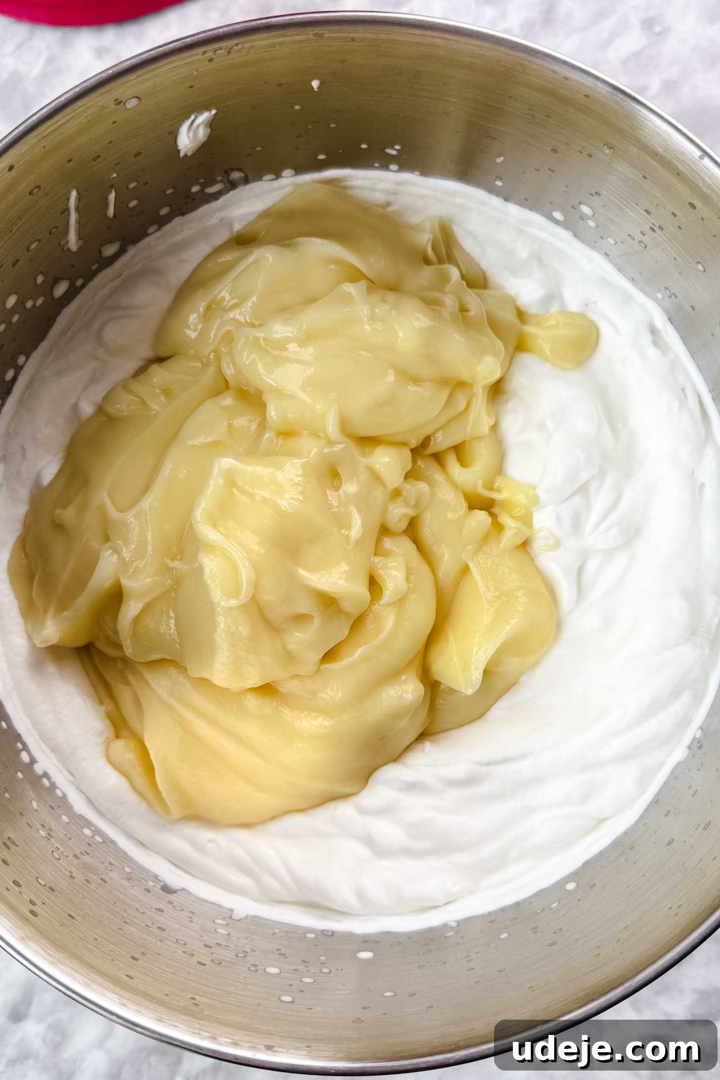 Magnolia Bakery's Irresistible Banana Pudding 8 Thick yellow pudding in the silver bowl of a stand mixer with whipped cream on a marble background.