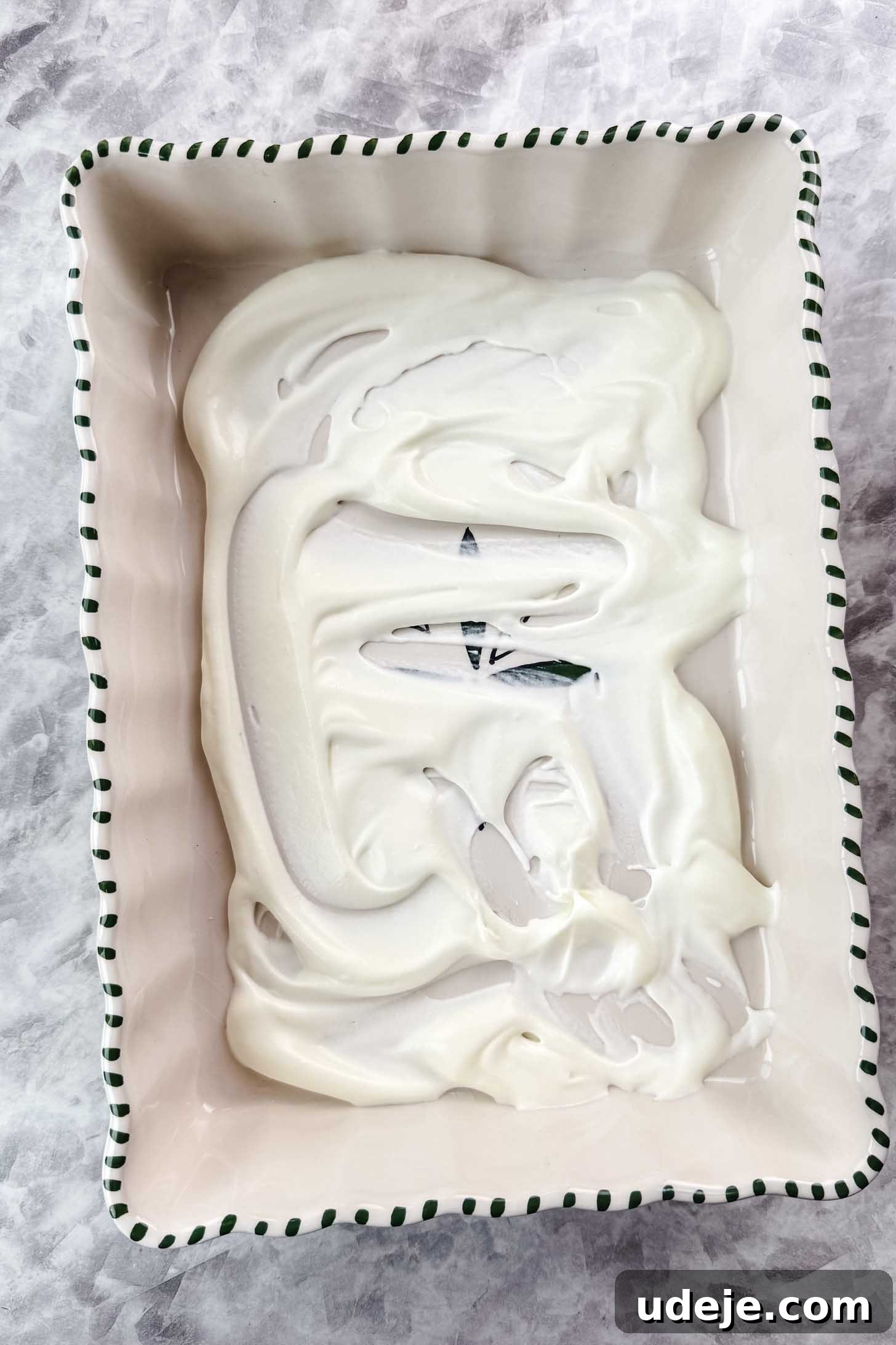 Magnolia Bakery's Irresistible Banana Pudding 10 White baking dish with green stripes on the edge with cream filling spread at the bottom on a marble background.
