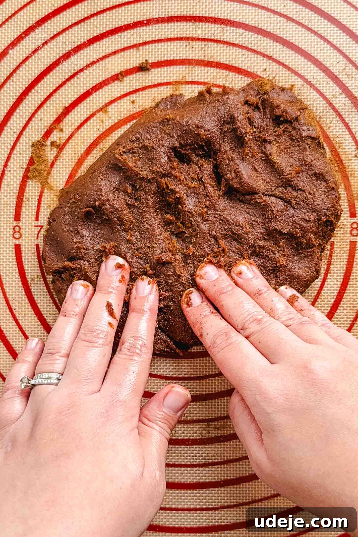 Iraqi Kleicha A Sweet Tradition with Date or Walnut 11 Two hands knead a mound of Kleicha chocolate cookie dough on a red and tan silicone baking mat with concentric circles and measurement markings.