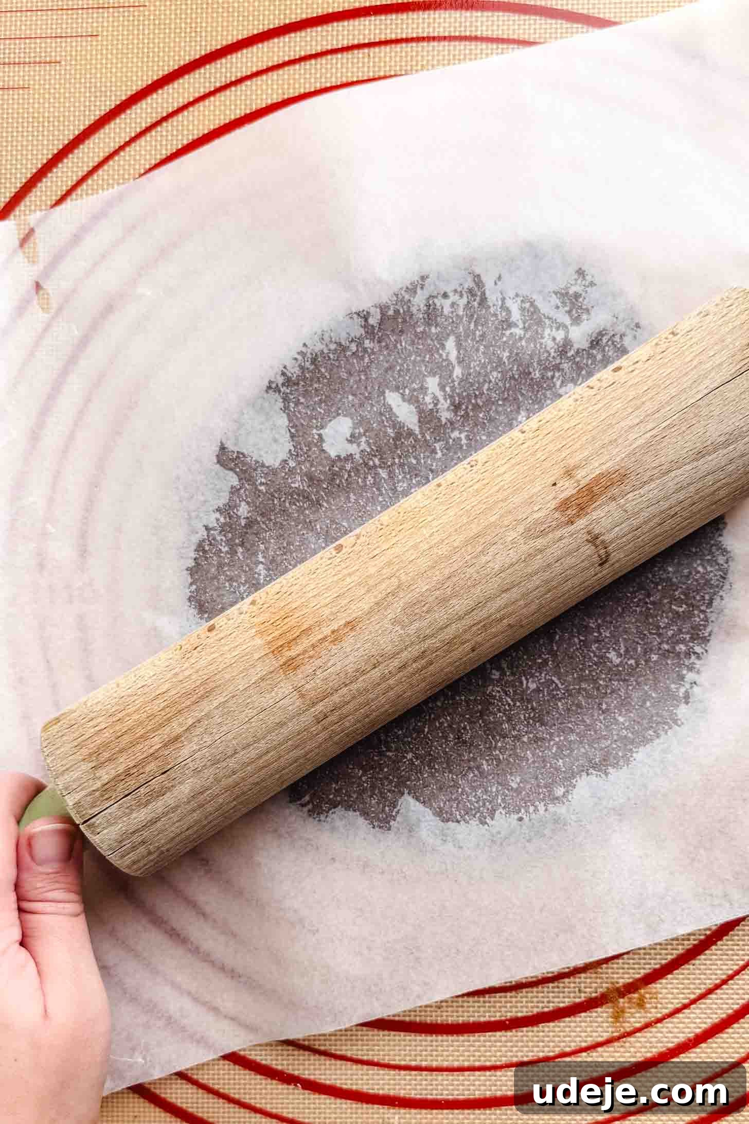 Iraqi Kleicha A Sweet Tradition with Date or Walnut 12 A hand holds a wooden rolling pin, flattening kleicha dough between two sheets of parchment paper on a red-rimmed silicone baking mat.