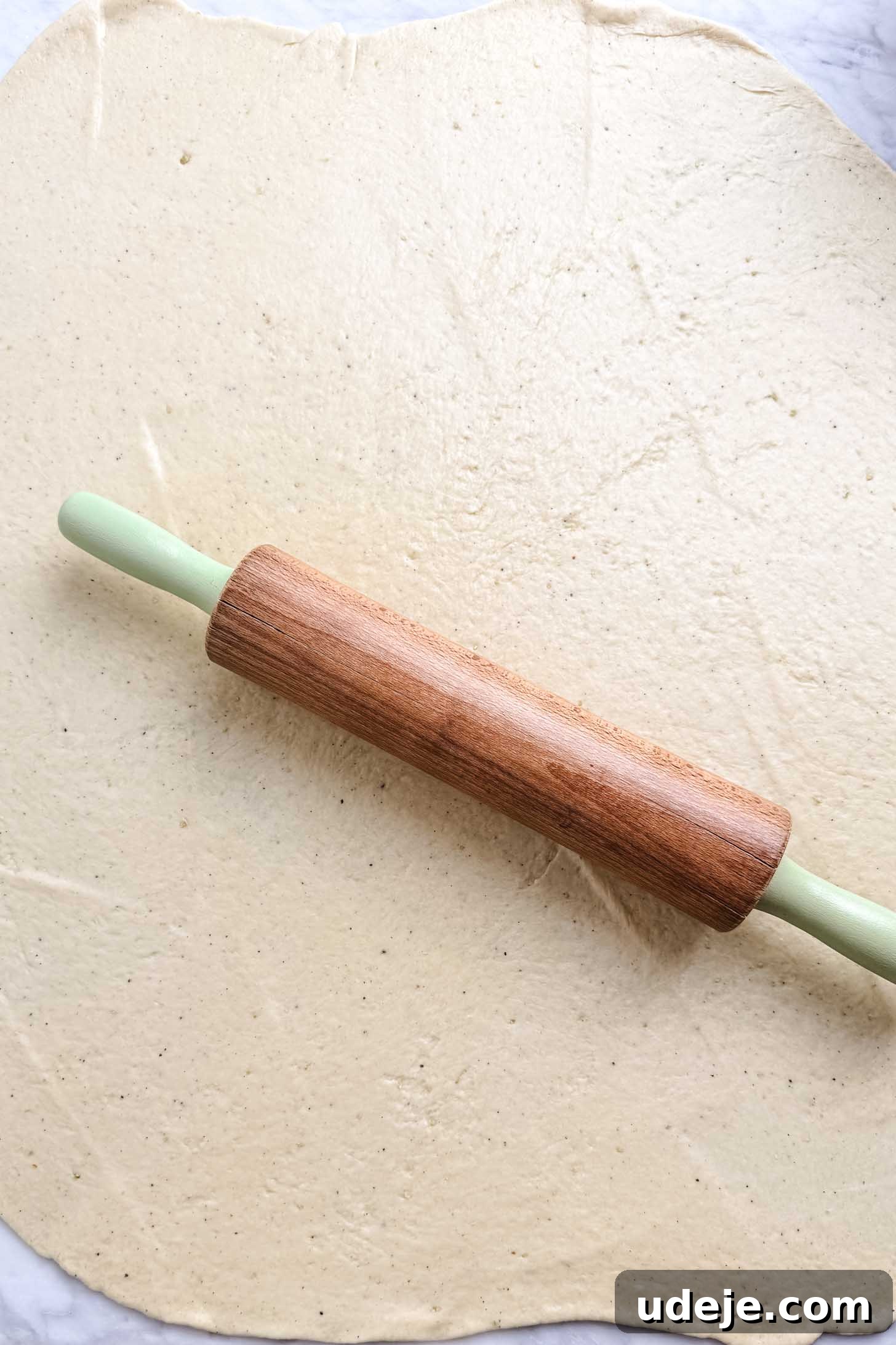 Iraqi Kleicha A Sweet Tradition with Date or Walnut 14 A wooden rolling pin with green handles rests on top of a large, evenly rolled sheet of dough—perfect for shaping traditional Kleicha—on a light-colored surface.