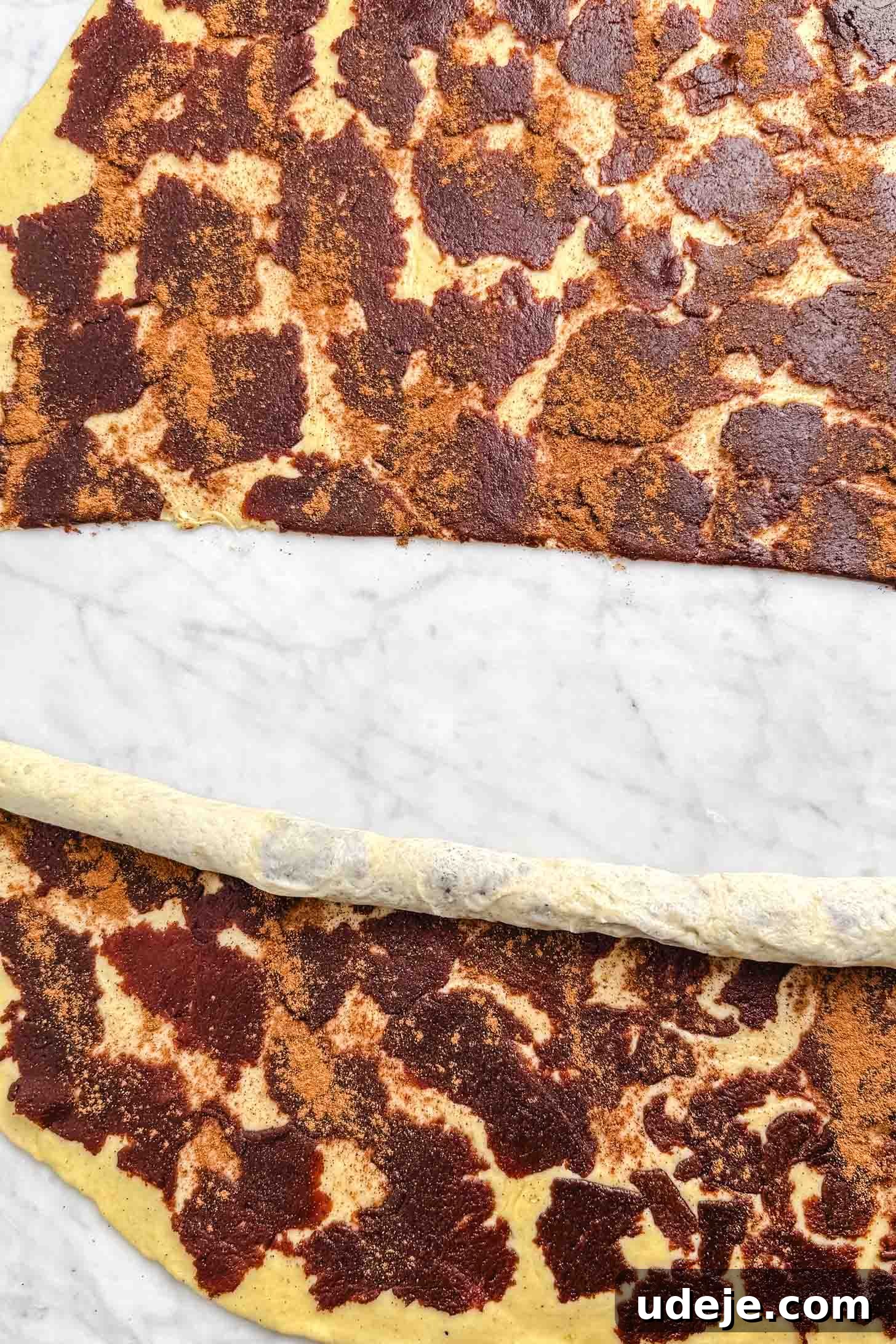 Iraqi Kleicha A Sweet Tradition with Date or Walnut 16 A sheet of dough spread with a brown cinnamon mixture is being rolled up on a marble surface, much like the traditional preparation of Kleicha, preparing to make delicious cinnamon rolls.