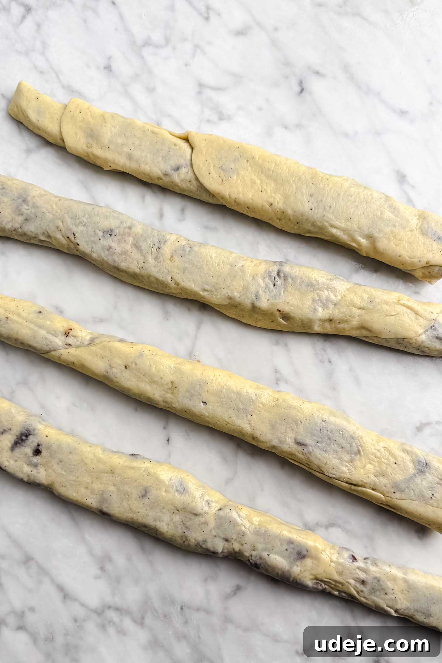 Iraqi Kleicha A Sweet Tradition with Date or Walnut 17 Four rolled strands of dough, reminiscent of traditional Kleicha, with visible chocolate pieces lie parallel on a white marble surface, ready for further shaping or baking preparation.