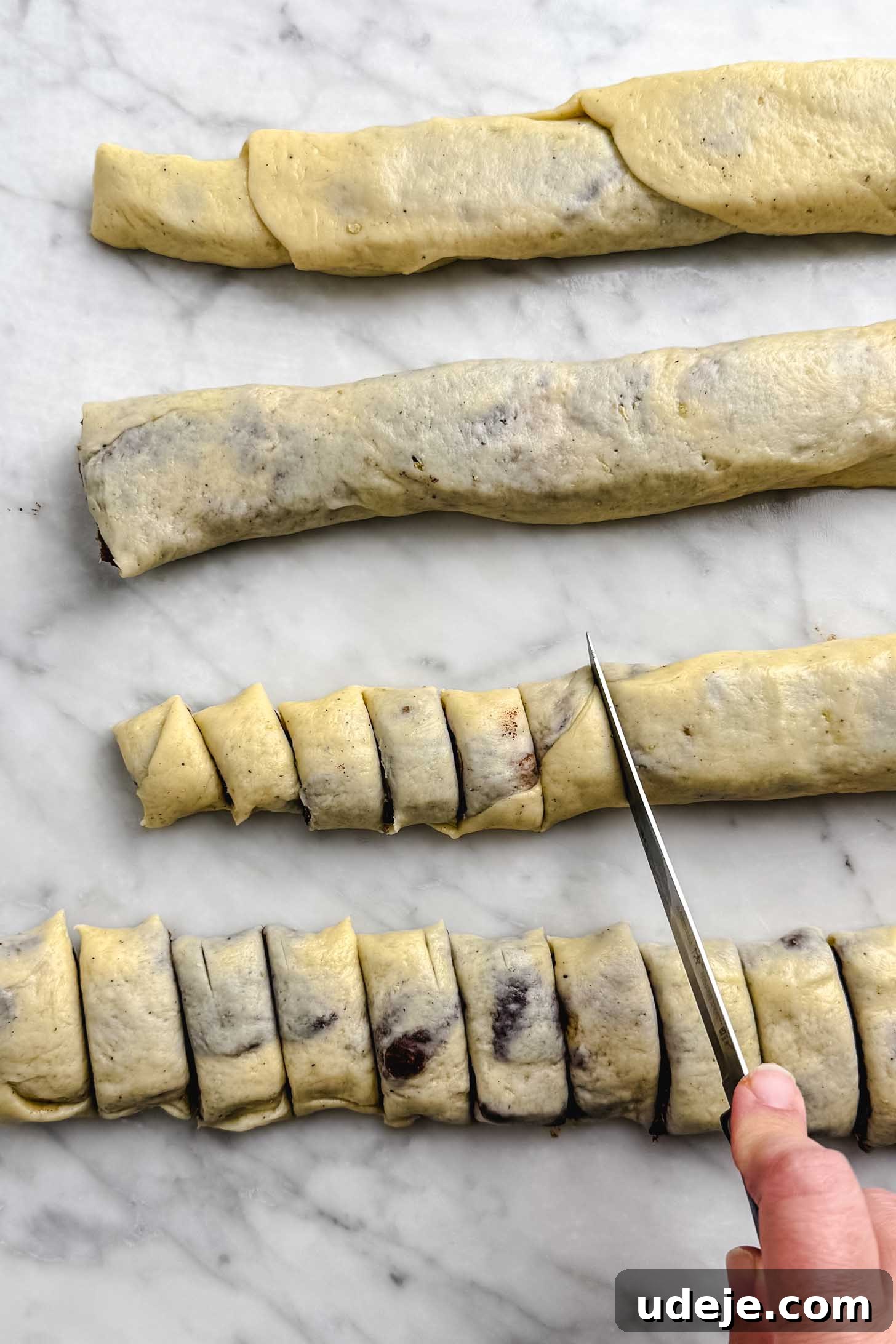 Iraqi Kleicha A Sweet Tradition with Date or Walnut 18 A hand uses a knife to cut rolled Kleicha dough with a dark filling into small pieces on a marble surface. Three uncut dough rolls are above the cut pieces.