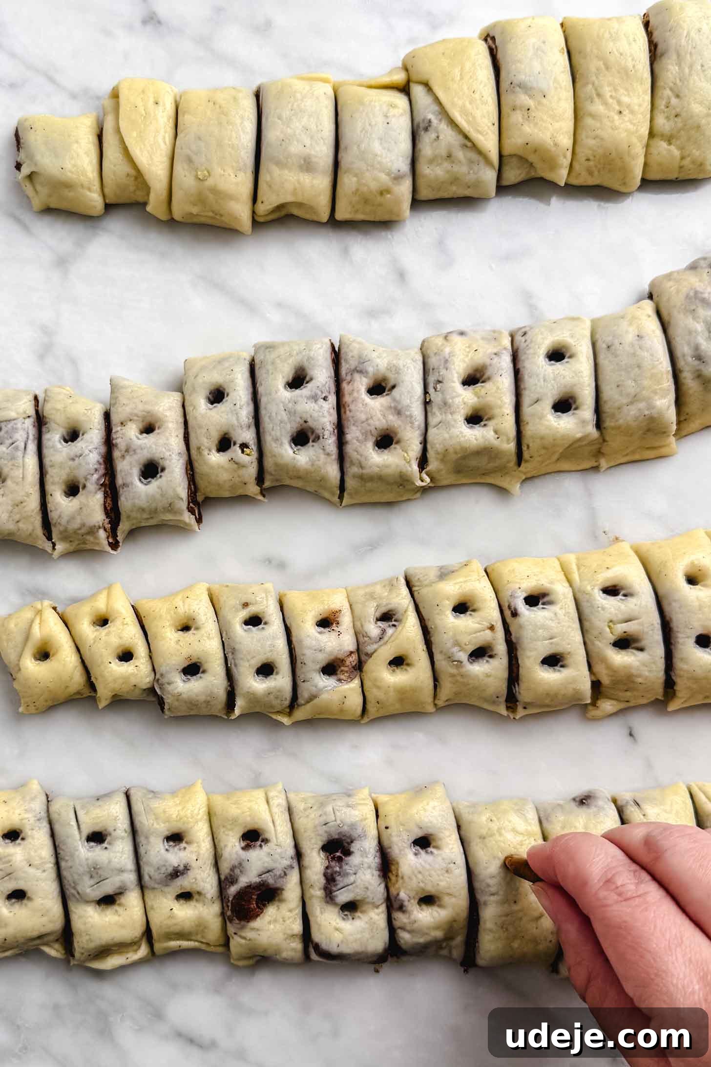 Iraqi Kleicha A Sweet Tradition with Date or Walnut 19 Strips of dough filled with chocolate, inspired by Kleicha, are cut into small pieces and arranged on a marble surface. Each piece has holes poked in the top, and a hand is poking holes in one piece.
