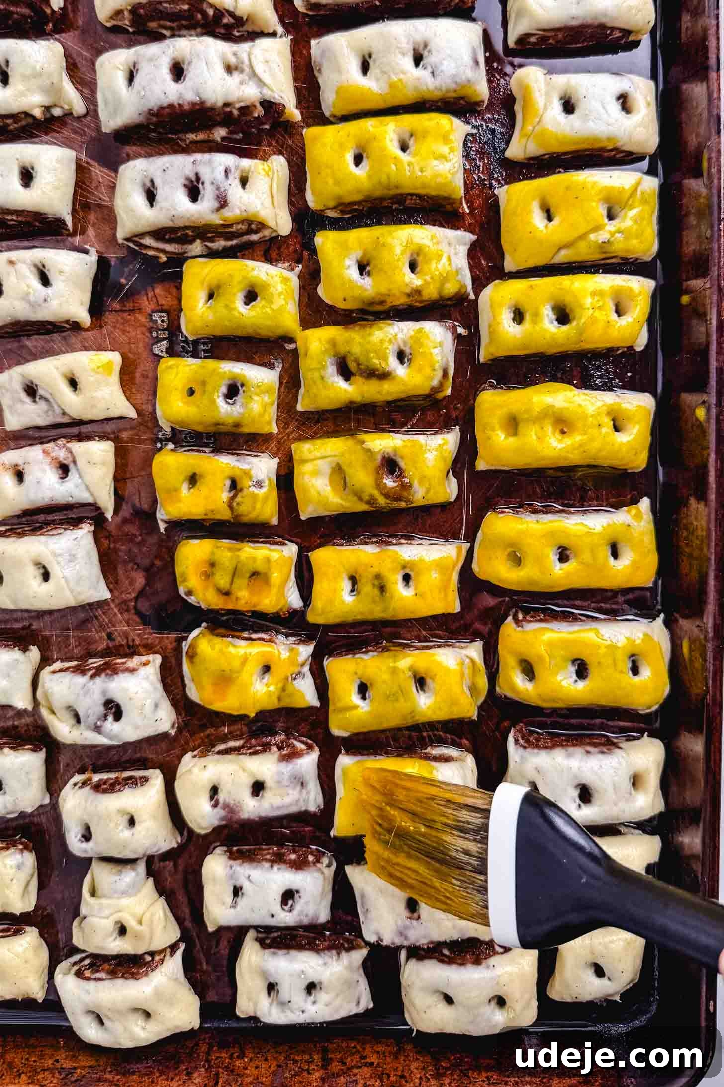Iraqi Kleicha A Sweet Tradition with Date or Walnut 20 Rows of unbaked Kleicha pastry rolls, some brushed with egg wash for a golden tint, are arranged on a baking sheet. A pastry brush gently applies egg wash to one roll in the corner.
