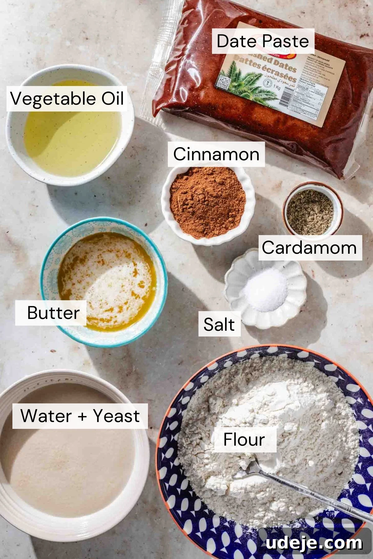 Iraqi Kleicha A Sweet Tradition with Date or Walnut 3 Bowls and containers holding labeled ingredients for baking Kleicha: vegetable oil, melted butter, date paste, cinnamon, cardamom, salt, water with yeast, and flour—all arranged on a light surface.