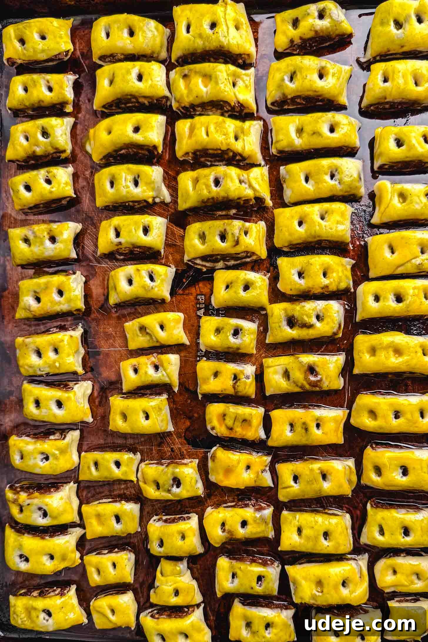Iraqi Kleicha A Sweet Tradition with Date or Walnut 21 Rows of rectangular, golden-brown Kleicha pastries with three small holes on top are arranged neatly on a baking tray. Each Kleicha shines with a glossy finish and is uniformly spaced, highlighting their perfect preparation.