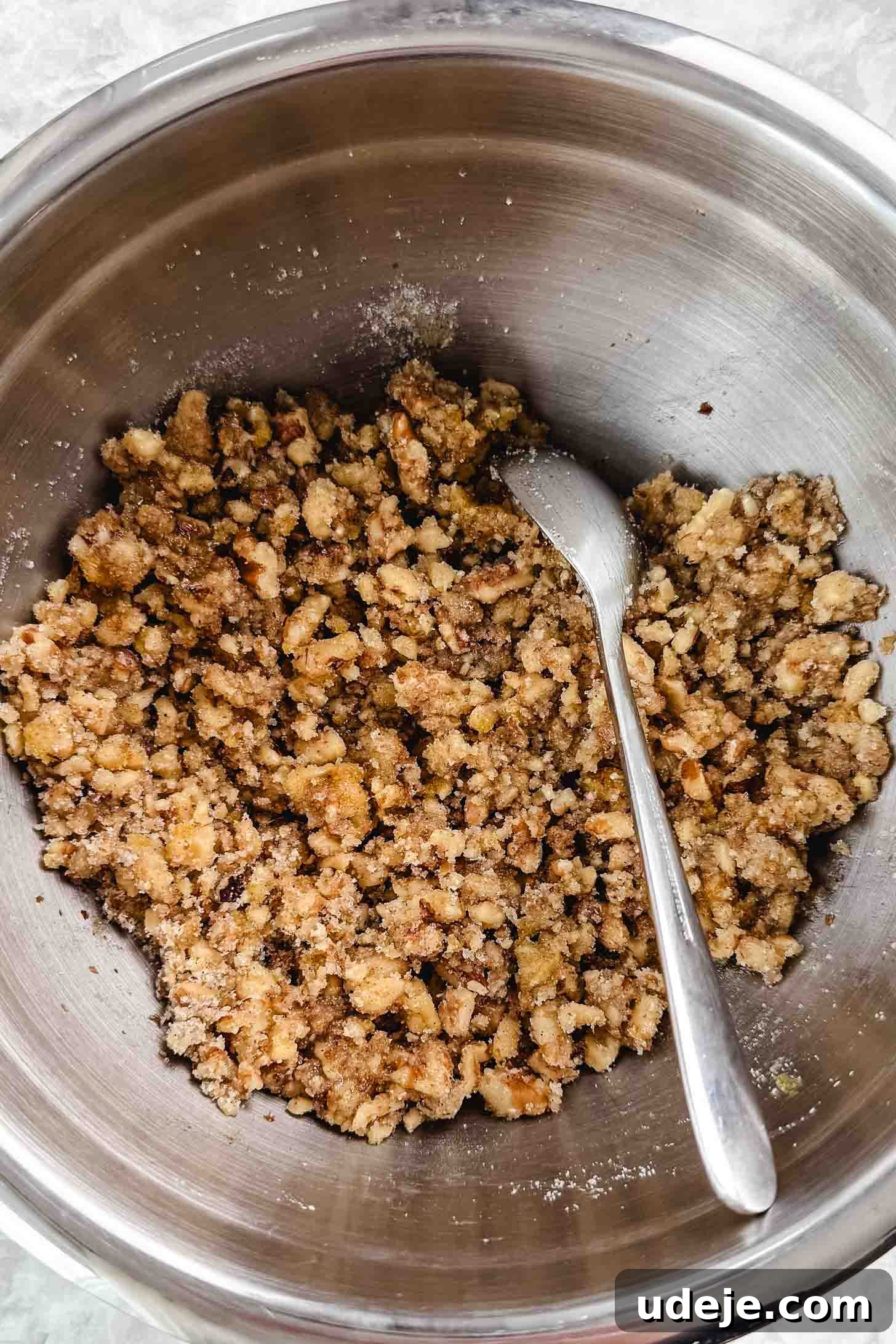 Iraqi Kleicha A Sweet Tradition with Date or Walnut 22 A metal mixing bowl filled with a crumbly mixture of brown sugar, flour, and chopped nuts for Kleicha, with a metal spoon resting on the side.
