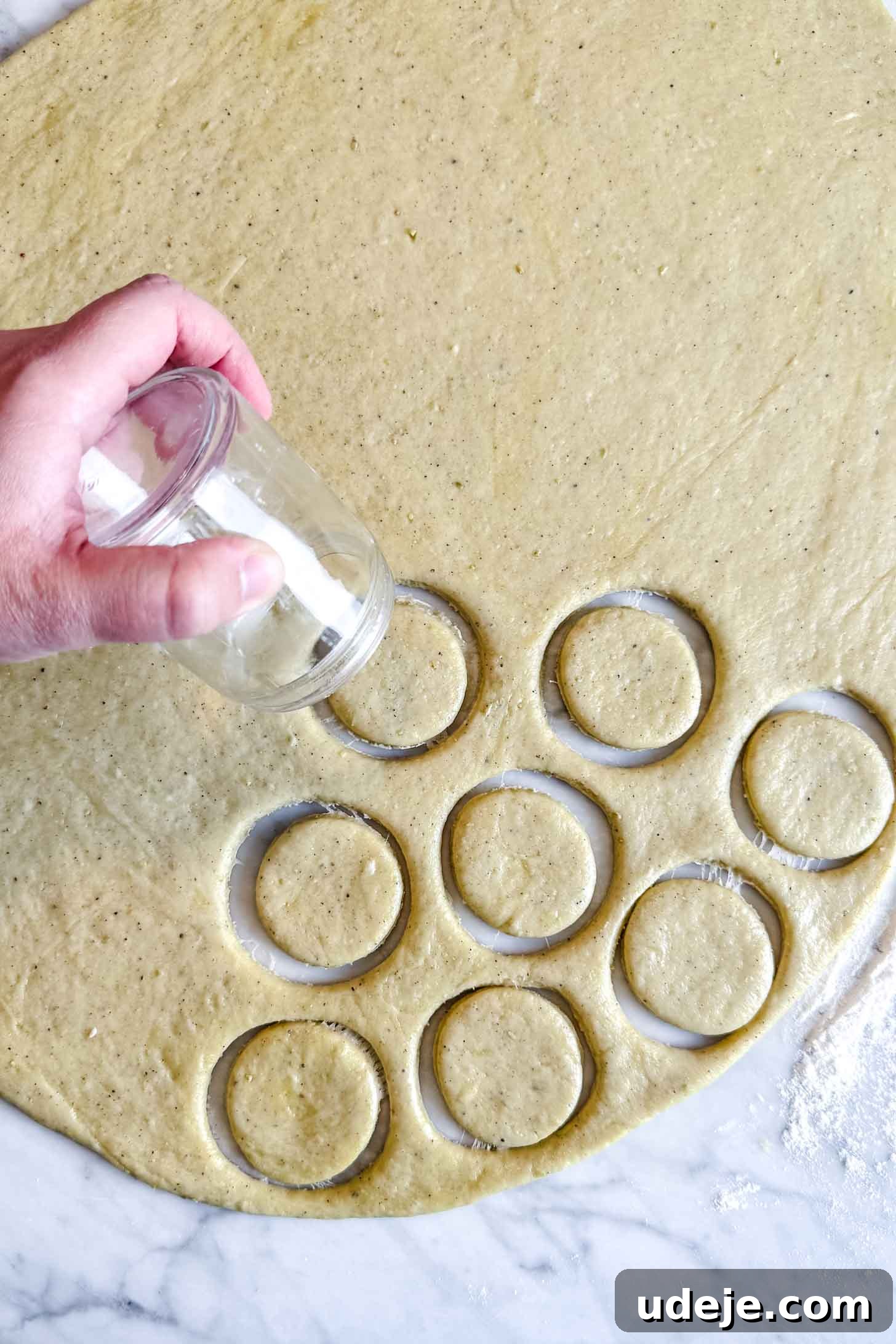 Iraqi Kleicha A Sweet Tradition with Date or Walnut 23 A hand uses a glass to cut circular shapes for Kleicha from rolled-out dough on a floured surface. Several dough circles are already cut out and visible next to the glass.