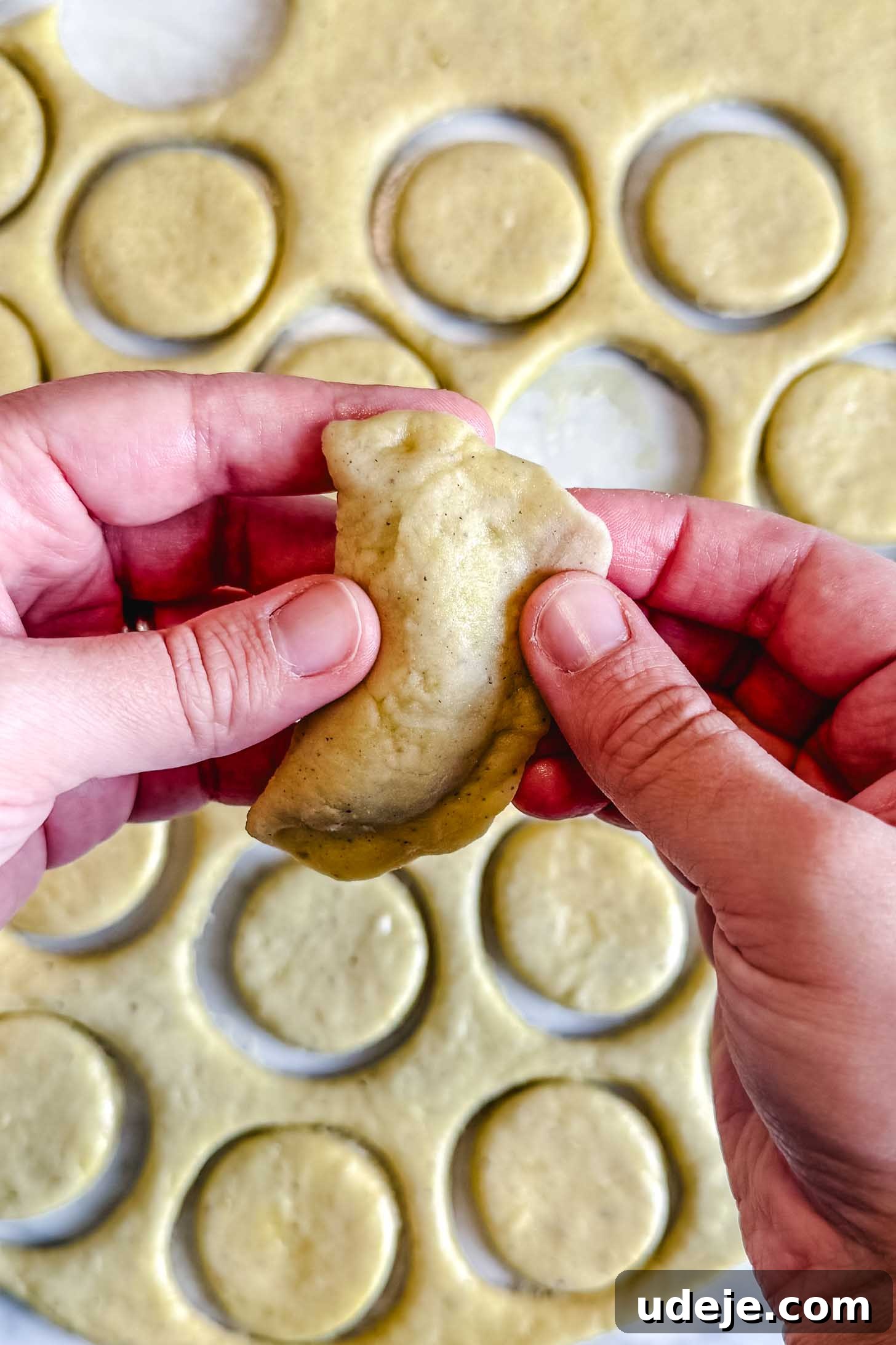 Iraqi Kleicha A Sweet Tradition with Date or Walnut 26 Hands shape a piece of dough into a half-moon, resembling the traditional Kleicha, with round dough cutouts and rolled dough visible on a work surface in the background.