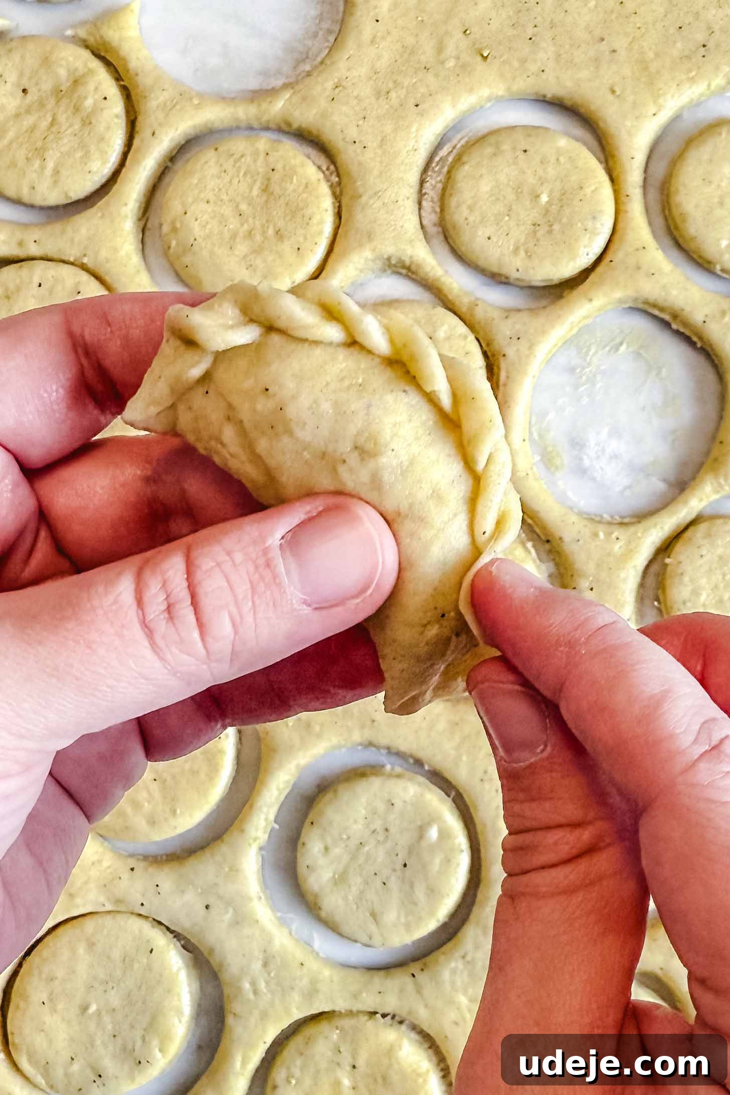 Iraqi Kleicha A Sweet Tradition with Date or Walnut 27 A person’s hands fold and pinch the edges of a round kleicha or pastry, with circles of dough and cutouts scattered on a floured surface in the background.