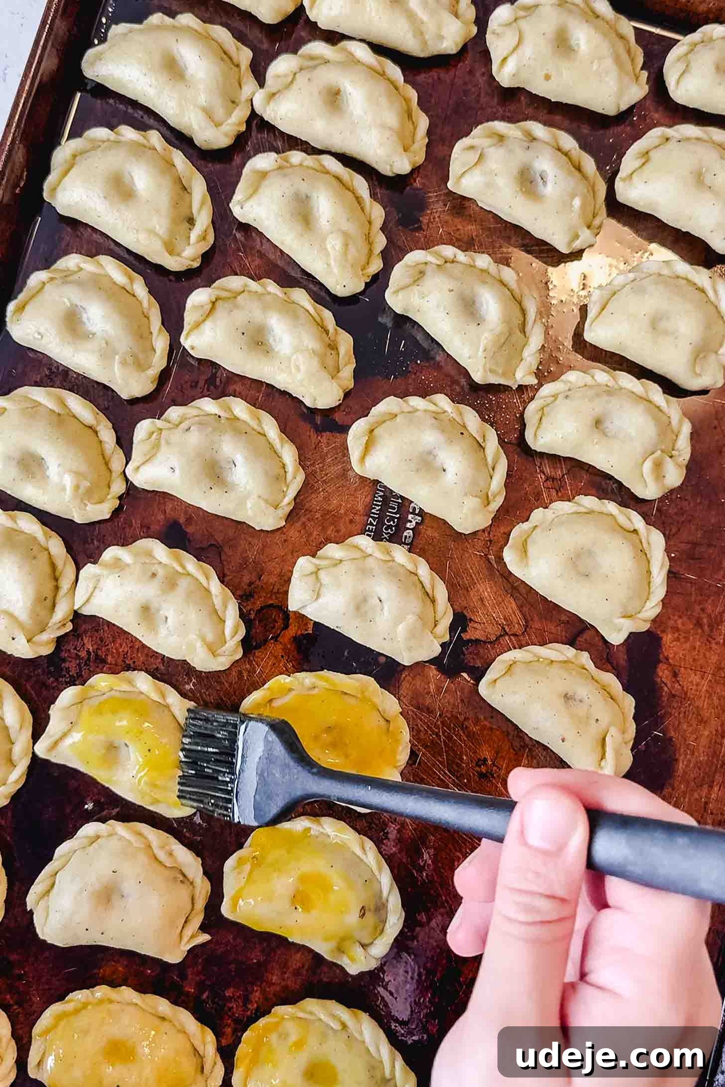 Iraqi Kleicha A Sweet Tradition with Date or Walnut 28 A hand uses a brush to apply egg wash to rows of uncooked, folded Kleicha dumplings arranged on a baking sheet.