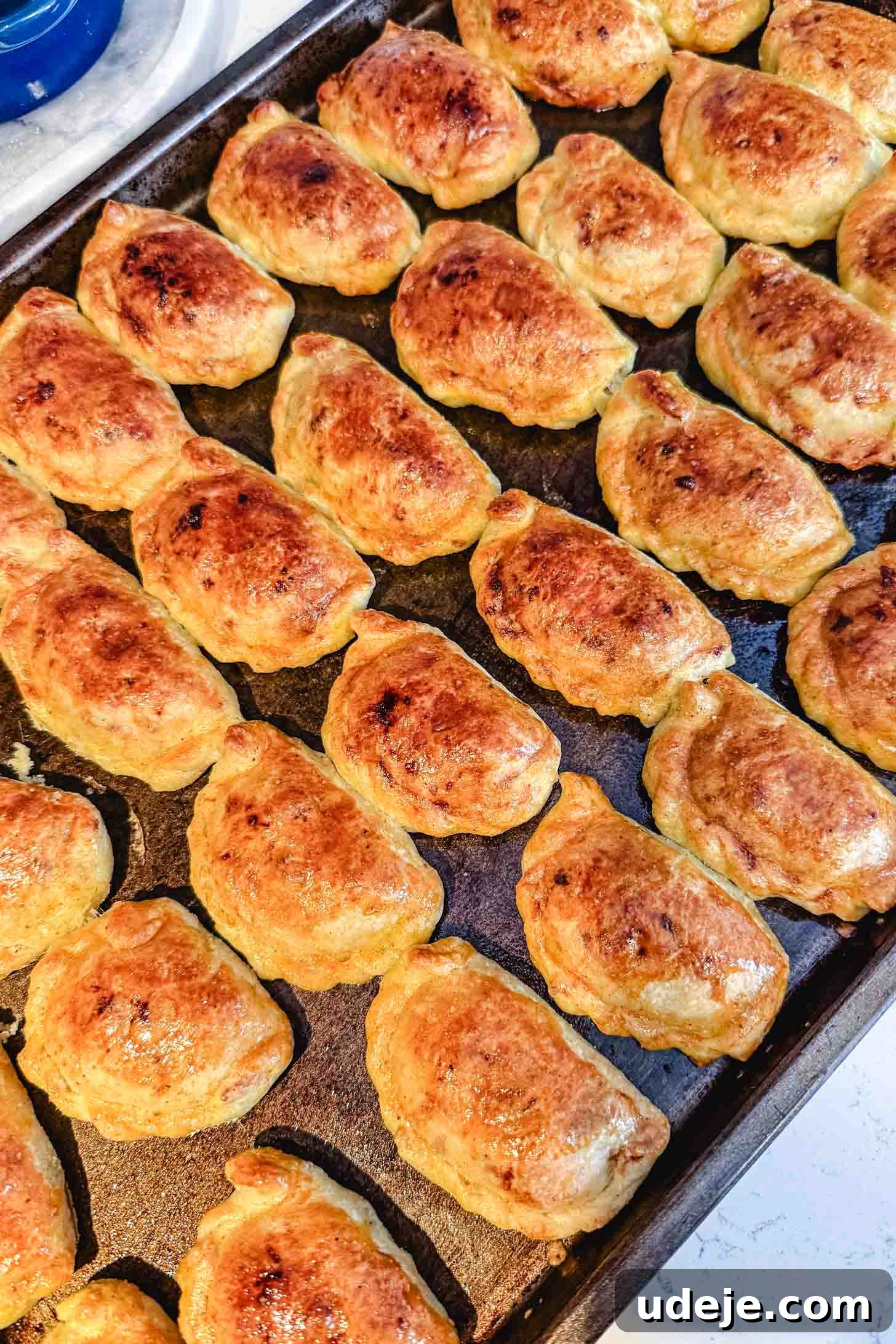 Iraqi Kleicha A Sweet Tradition with Date or Walnut 29 A baking tray filled with rows of golden-brown, freshly baked stuffed pastries—likely kleicha hand pies or empanadas—arranged closely together.