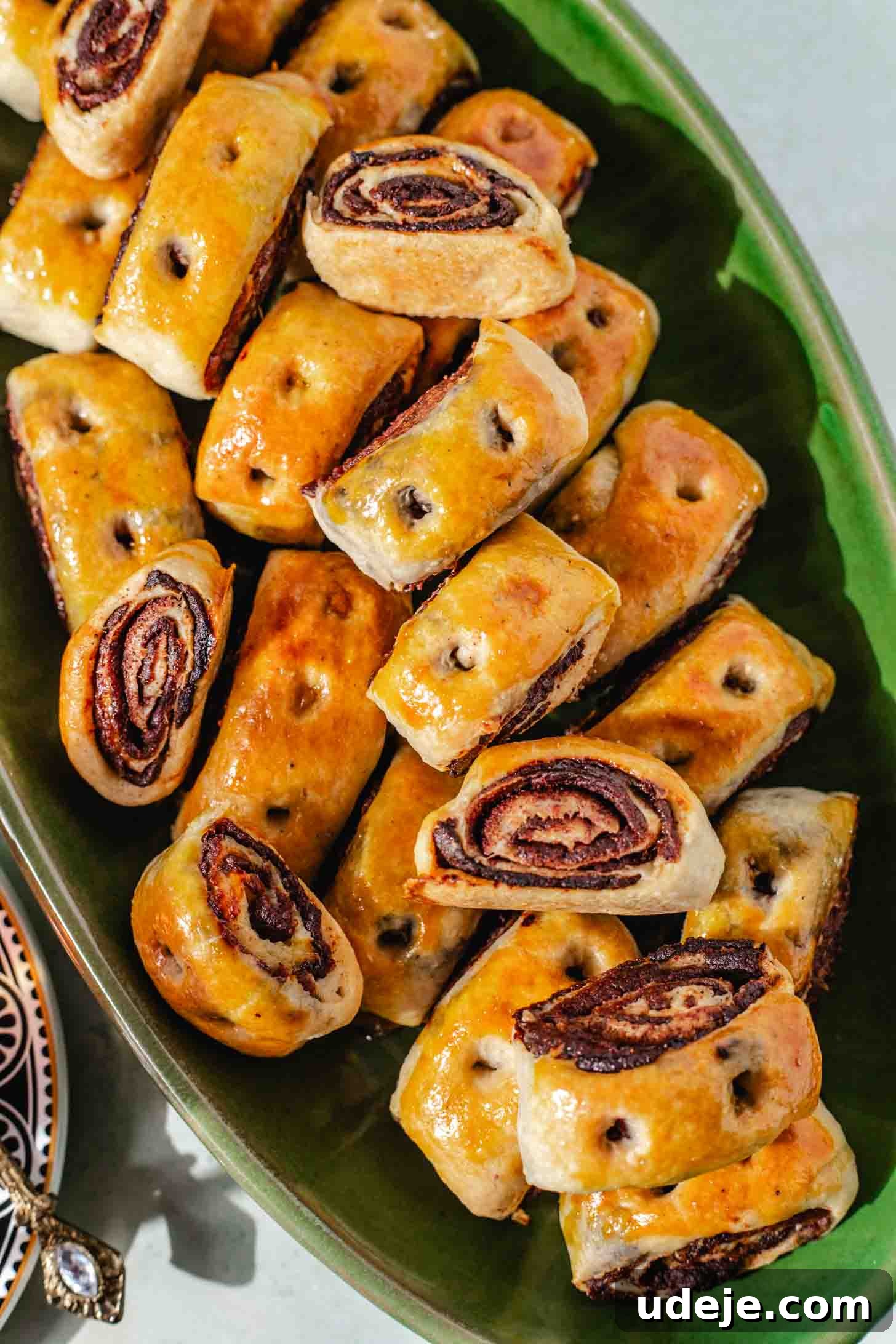 Iraqi Kleicha A Sweet Tradition with Date or Walnut 30 A green plate filled with golden-brown, rolled Kleicha pastries featuring visible chocolate or poppy seed swirls inside, arranged in neat, overlapping rows.