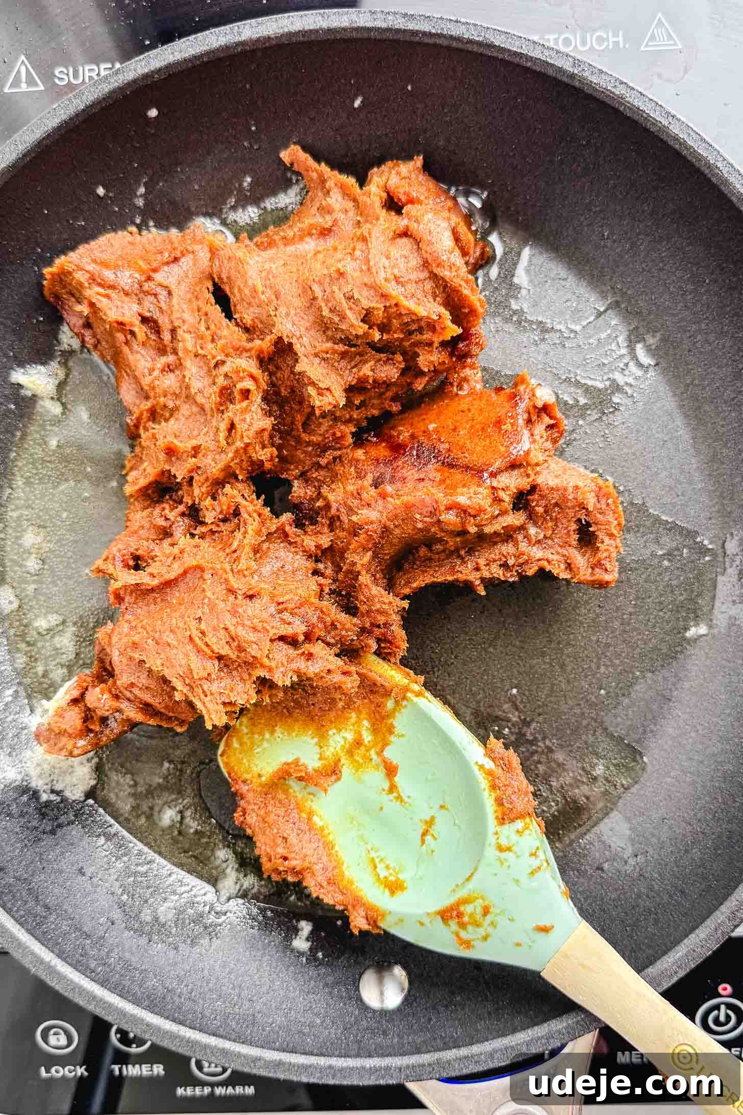 Iraqi Kleicha A Sweet Tradition with Date or Walnut 9 A green spatula stirs a thick, reddish-brown Kleicha paste in a black nonstick pan on an induction cooktop, with melted oil visible around the paste.