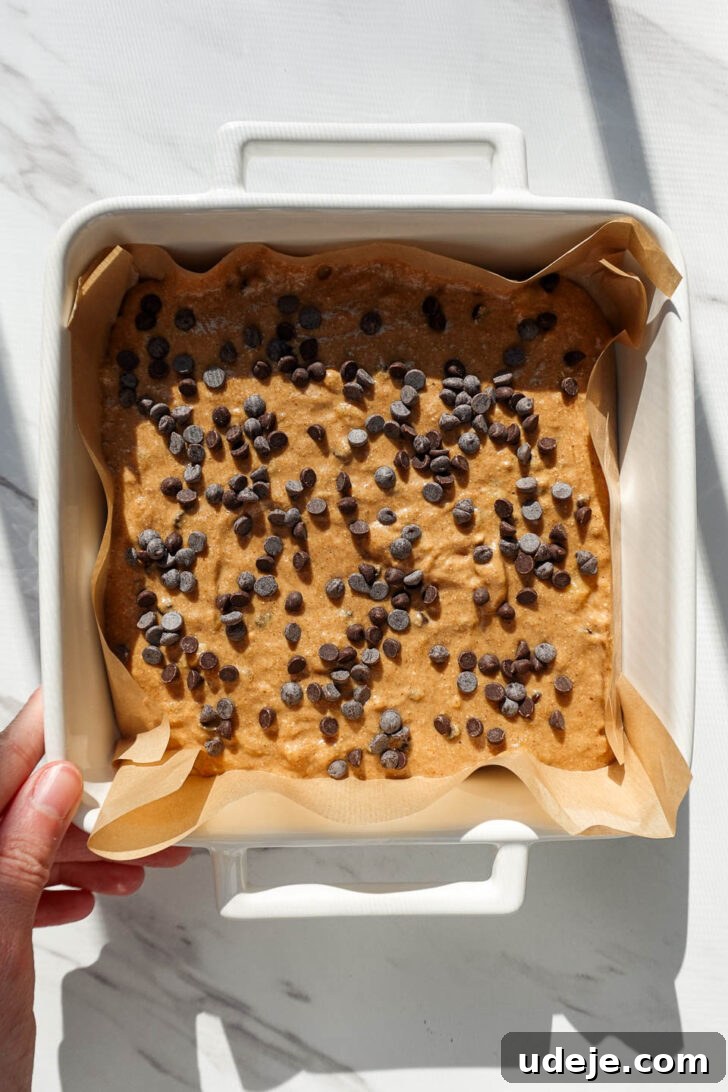 Vegan Gluten Free Chocolate Chip Banana Dream Bars 8 Vegan banana bars batter spread evenly in a parchment-lined square baking pan, with mini chocolate chips sprinkled generously on top before baking.