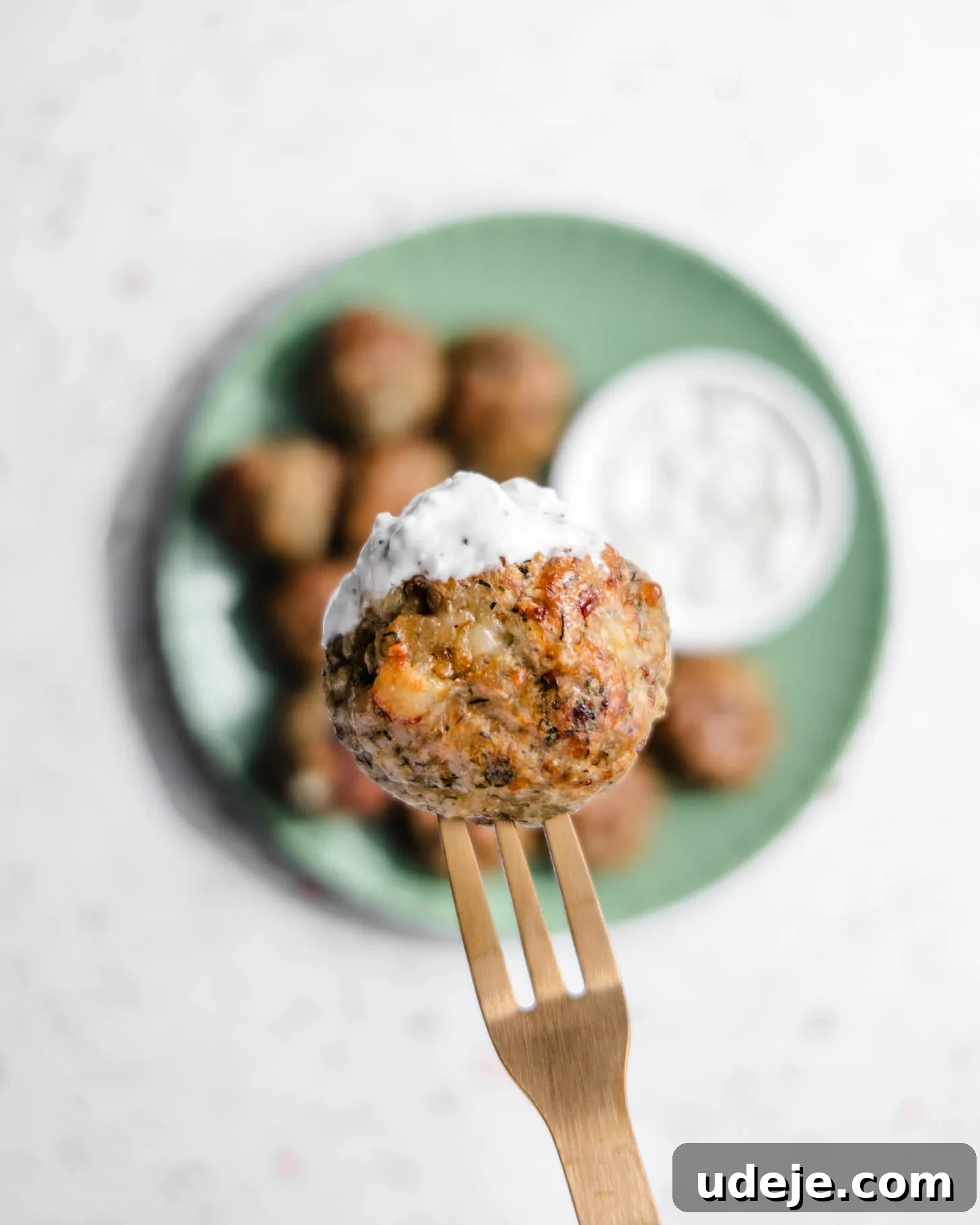 Baked Greek Chicken Meatballs with Zesty Tzatziki 11 A close-up shot capturing a single, perfectly baked Greek chicken meatball perched on a fork, glistening with a generous dollop of creamy white tzatziki sauce. In the softly blurred background, a green plate holds more enticing meatballs and a small bowl of the delicious dipping sauce, creating an irresistible visual of a Mediterranean feast.