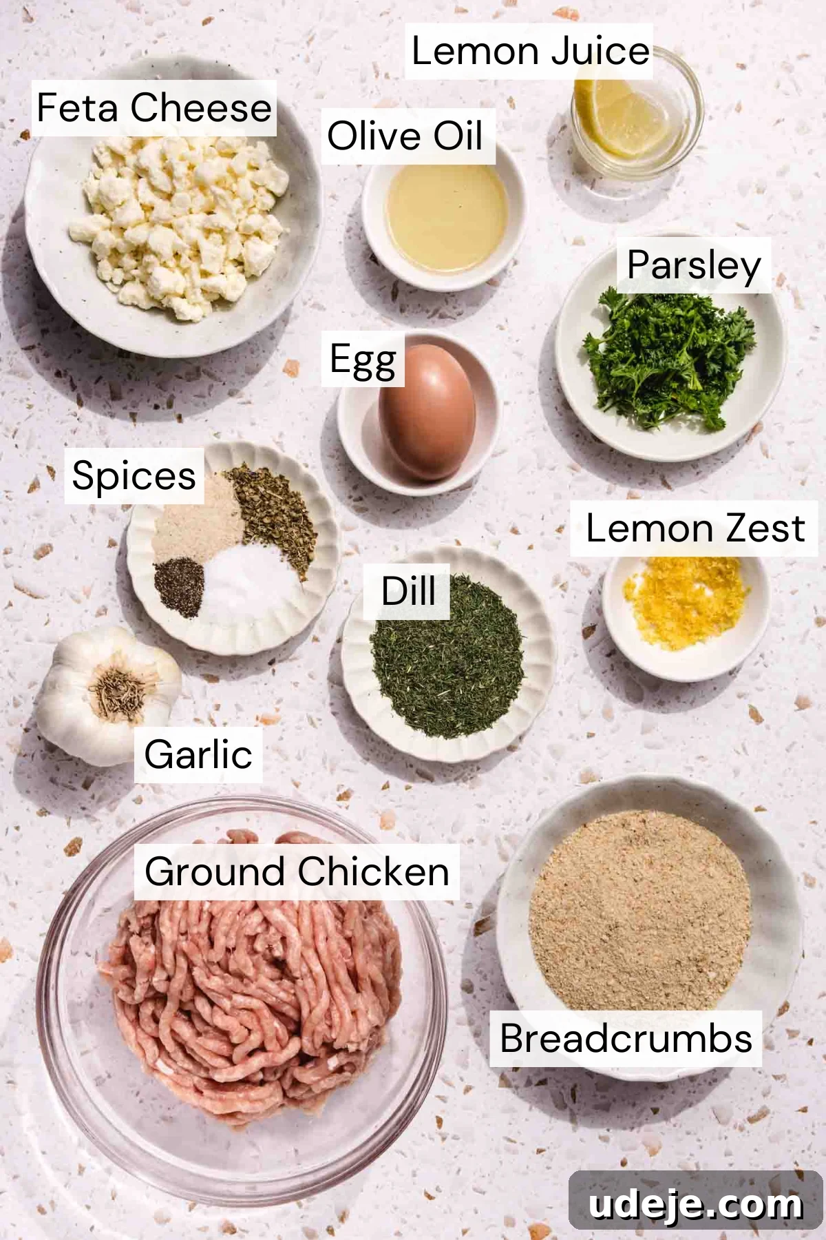 Baked Greek Chicken Meatballs with Zesty Tzatziki 3 A flat lay arrangement showcasing labeled ingredients for the Greek chicken meatballs. Bowls contain crumbled feta cheese, golden olive oil, fresh lemon juice, vibrant chopped parsley, a beaten egg, an assortment of spices, aromatic fresh dill, bright lemon zest, minced garlic, lean ground chicken, and dry breadcrumbs, all neatly presented on a light, textured surface, ready for mixing.