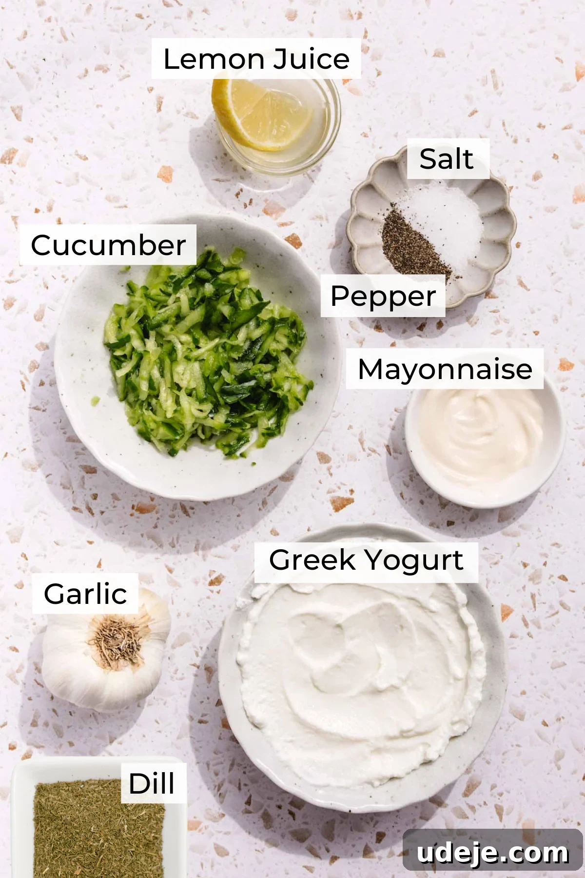 Baked Greek Chicken Meatballs with Zesty Tzatziki 4 An organized display of ingredients for homemade tzatziki sauce on a clean countertop. Bowls hold fresh lemon juice, fine salt, coarse black pepper, finely grated cucumber, creamy mayonnaise, rich Greek yogurt, pungent minced garlic, and aromatic dried dill, all prepared for mixing to create the perfect dipping sauce.