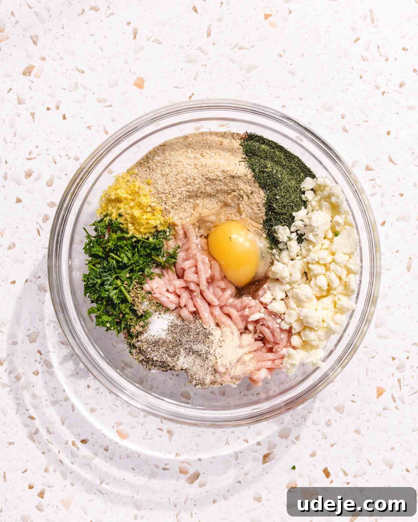 Baked Greek Chicken Meatballs with Zesty Tzatziki 5 A clear glass mixing bowl containing all the individual components for the Greek chicken meatballs, arranged in distinct sections: ground chicken, an egg, breadcrumbs, vibrant chopped herbs (dill and parsley), crumbled feta cheese, and various seasonings, all resting on a white speckled countertop, ready to be combined.