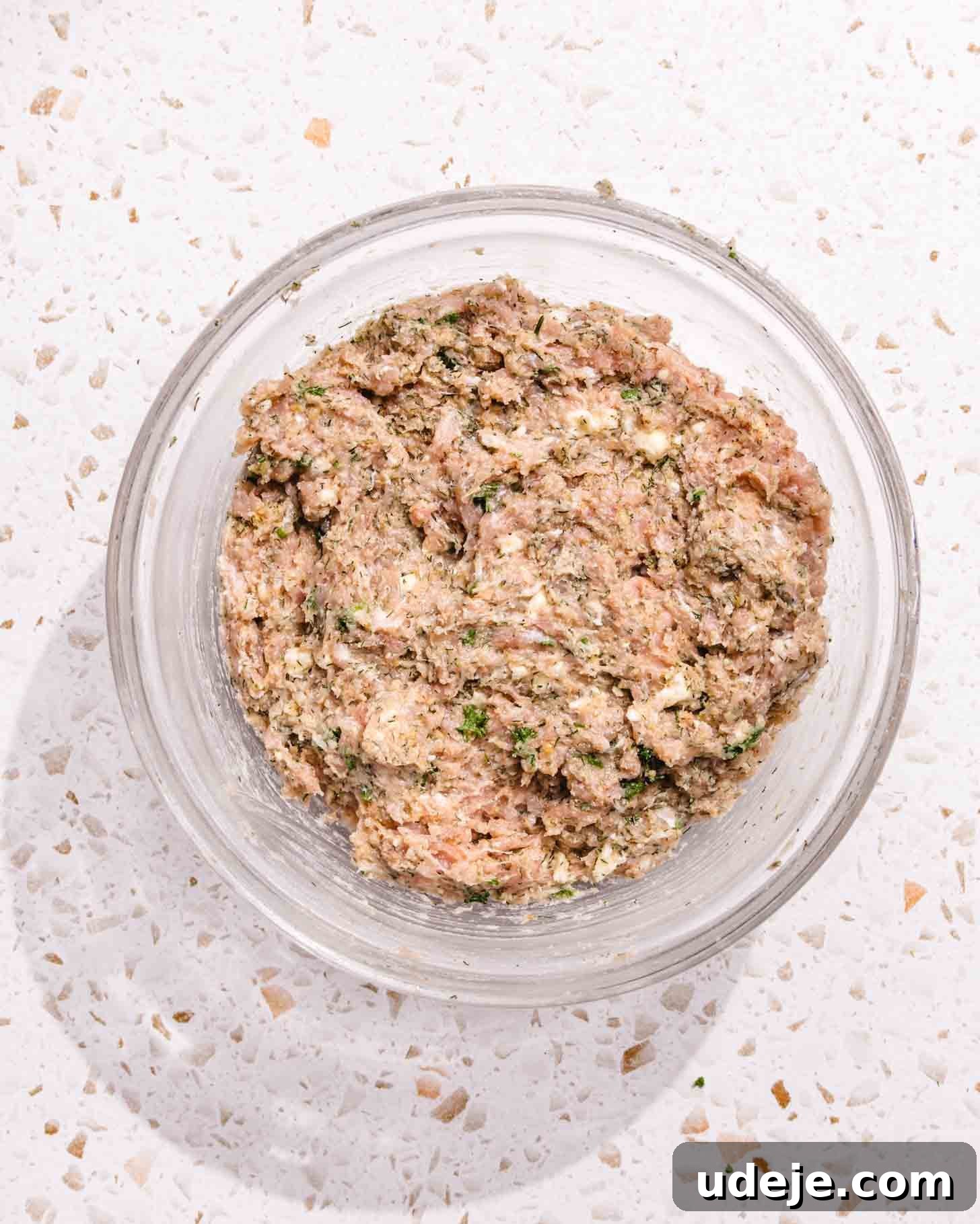 Baked Greek Chicken Meatballs with Zesty Tzatziki 6 A transparent glass bowl filled with the thoroughly mixed ground chicken mixture, showing a cohesive blend of meat, herbs, breadcrumbs, and feta, indicating it's ready for shaping. The bowl sits on a white speckled countertop.