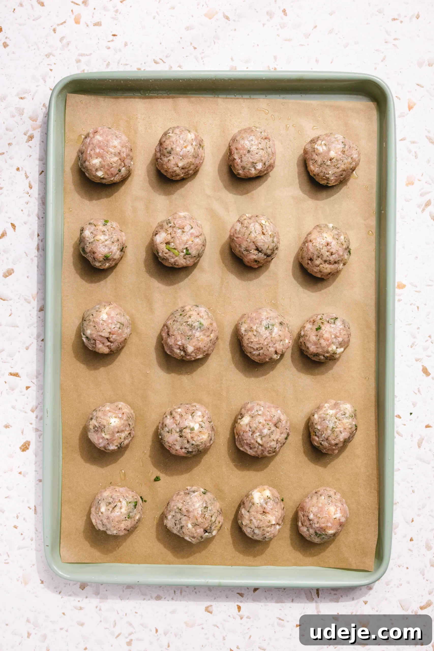 Baked Greek Chicken Meatballs with Zesty Tzatziki 7 A large green baking sheet, lined with rustic brown parchment paper, holding twenty uniformly shaped, uncooked Greek chicken meatballs. Each meatball is evenly spaced, showcasing flecks of green herbs and white feta, resting on a white, speckled kitchen countertop, ready for baking.