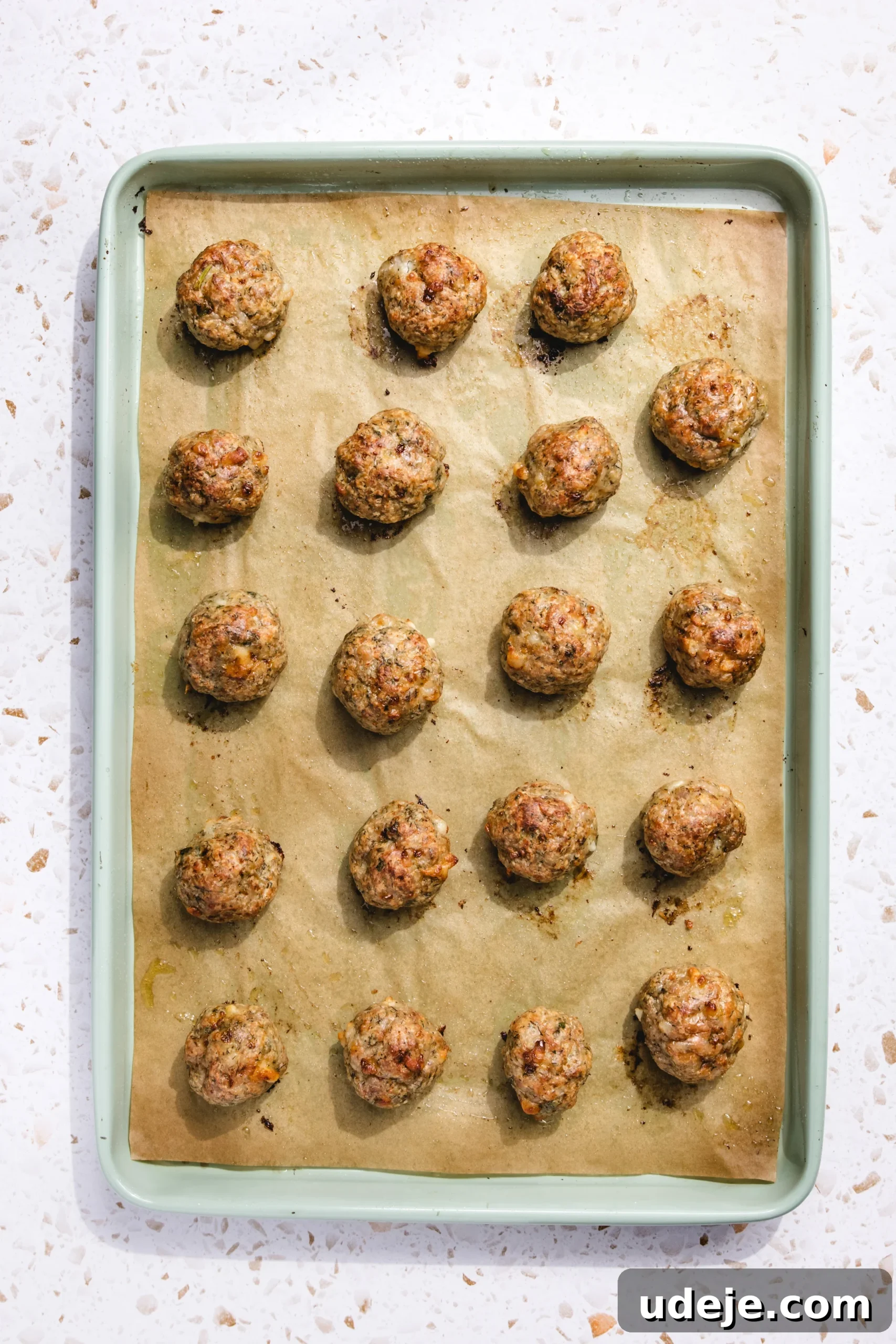 Baked Greek Chicken Meatballs with Zesty Tzatziki 8 A vibrant green baking tray, thoughtfully lined with brown parchment paper, now holds twenty perfectly baked Greek chicken meatballs. They exhibit a beautiful golden-brown exterior, evenly spaced across the tray, and are presented on a white speckled kitchen surface, ready to be served.