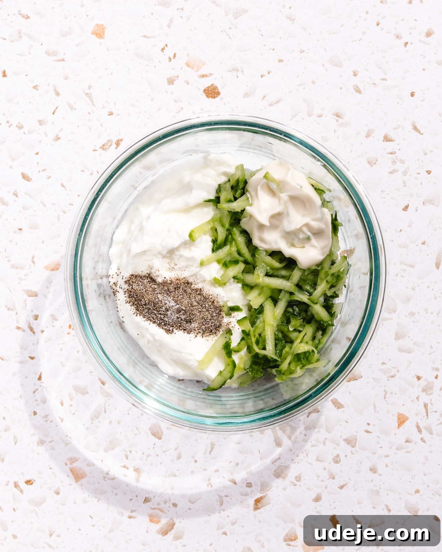 Baked Greek Chicken Meatballs with Zesty Tzatziki 9 A clear glass bowl on a speckled countertop containing the unmixed components of tzatziki: creamy Greek yogurt, finely shredded cucumber, a dollop of mayonnaise, and a sprinkle of ground black pepper, awaiting a gentle stir to transform into the classic sauce.