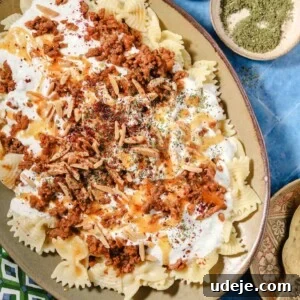 A beautiful platter of bowtie pasta topped with seasoned ground meat, a creamy white garlic yogurt sauce, delicate slivered almonds, fresh herbs, and aromatic spices, with small bowls of salt and dried herbs on the side for garnishing.