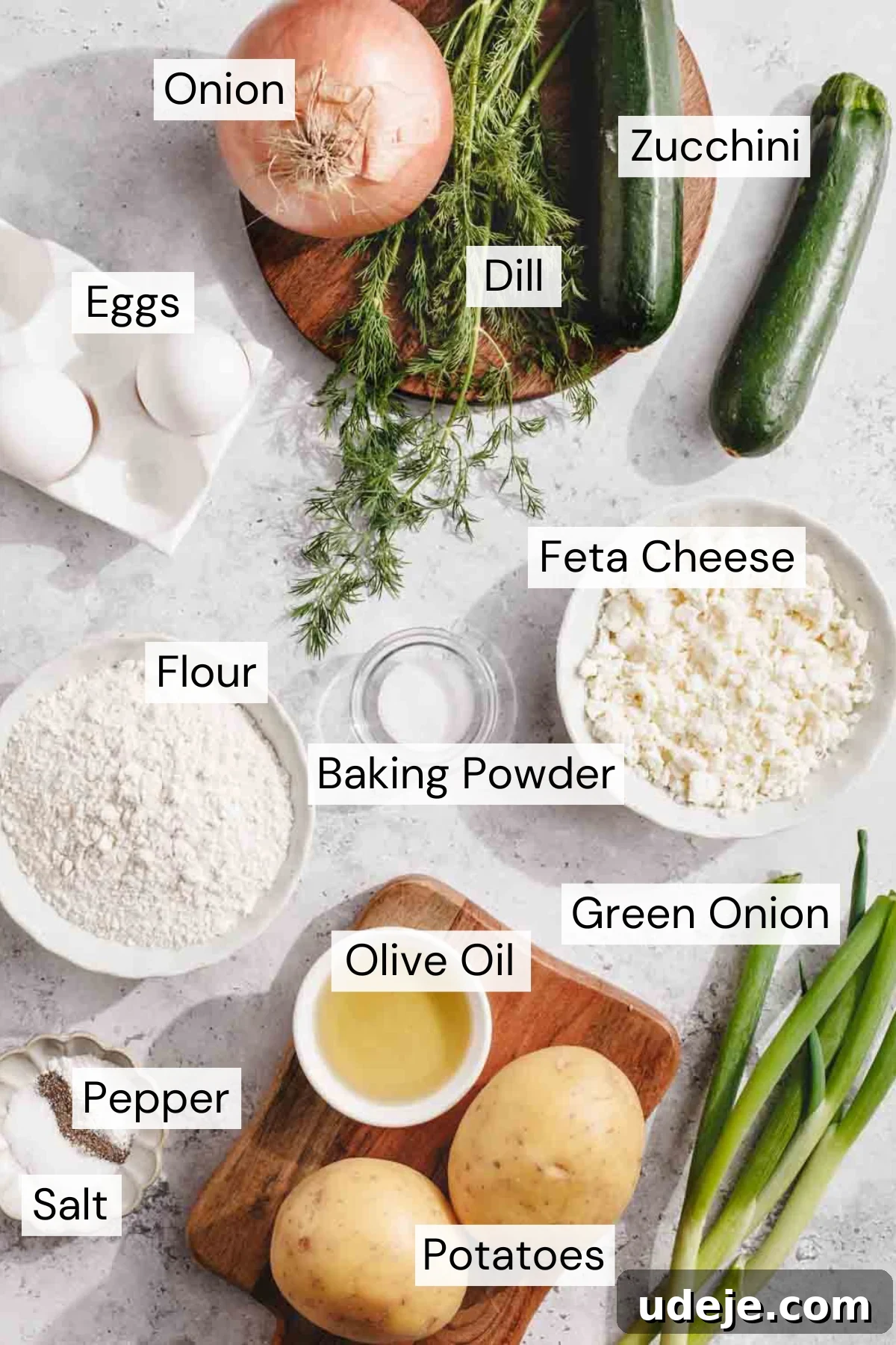 Crispy Turkish Zucchini Feta Fritters 3 Top-down view of various labeled ingredients on a light surface, including onion, zucchini, eggs, dill, flour, feta cheese, baking powder, green onion, olive oil, potatoes, salt, and pepper.