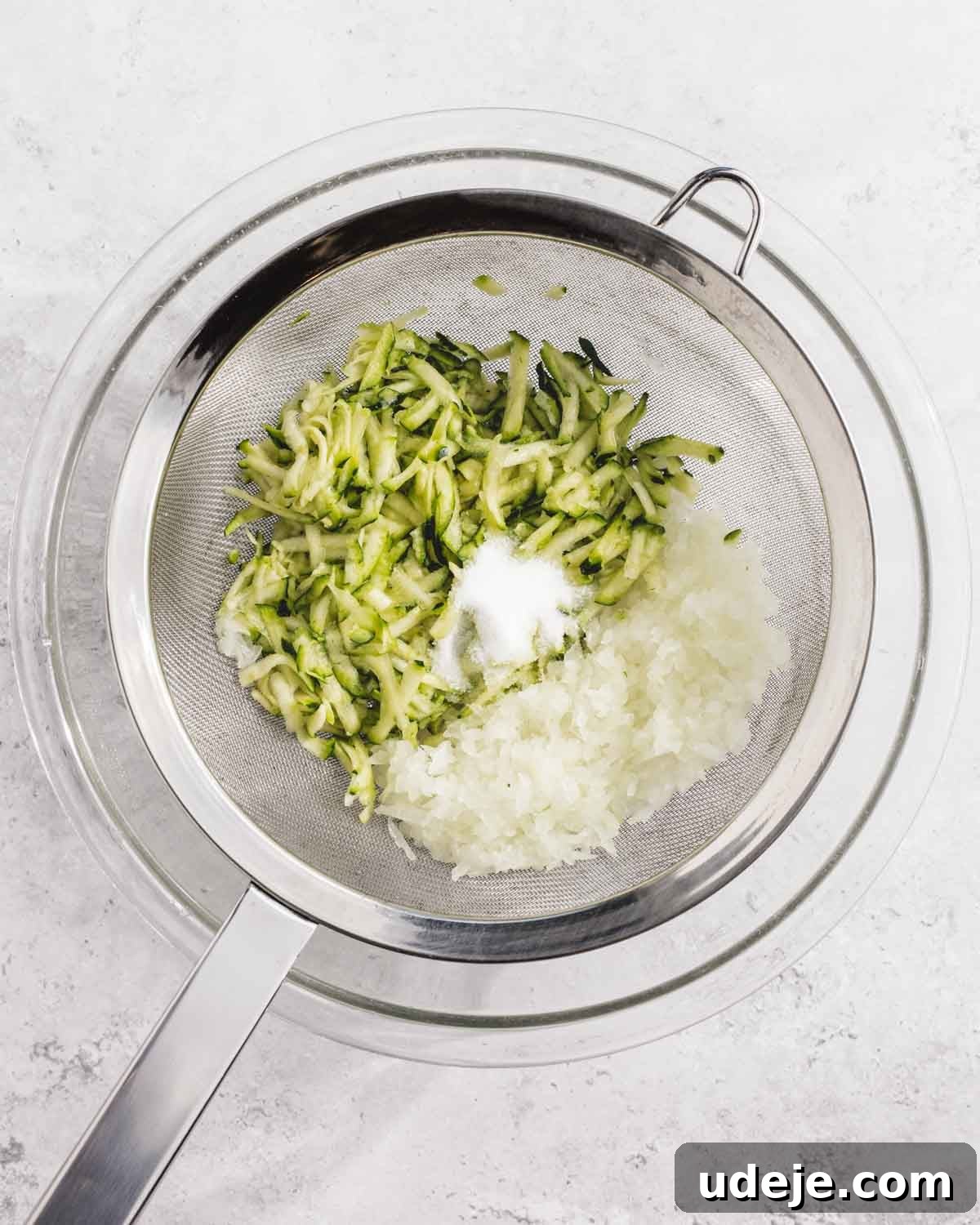 Crispy Turkish Zucchini Feta Fritters 4 A metal strainer over a glass bowl contains shredded zucchini, grated onion, and a small pile of salt. The mixture is set against a light, textured background.