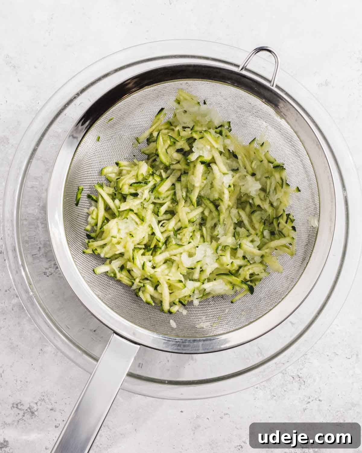 Crispy Turkish Zucchini Feta Fritters 5 A metal strainer with shredded zucchini rests over a glass bowl on a white surface. The zucchini appears to be draining or drying.