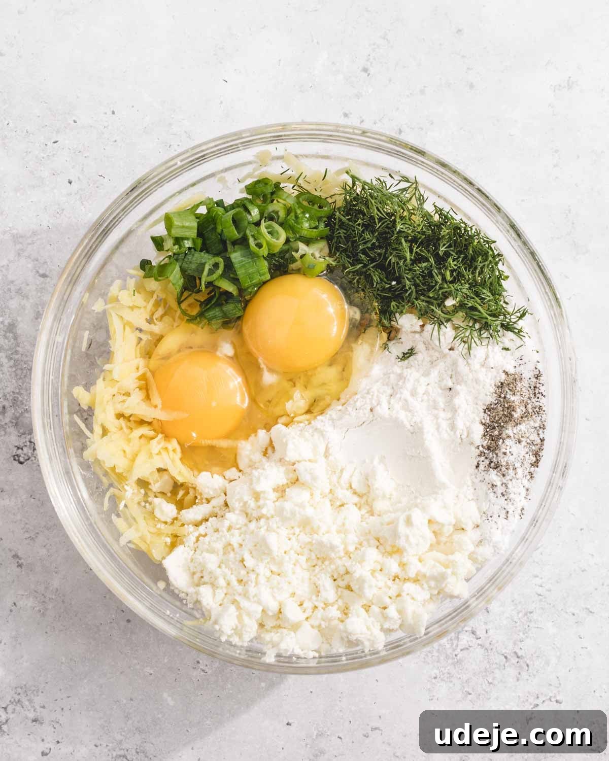 Crispy Turkish Zucchini Feta Fritters 6 A glass bowl containing eggs, shredded cheese, chopped green onions, fresh dill, flour, feta cheese, and ground black pepper, all arranged separately before mixing.