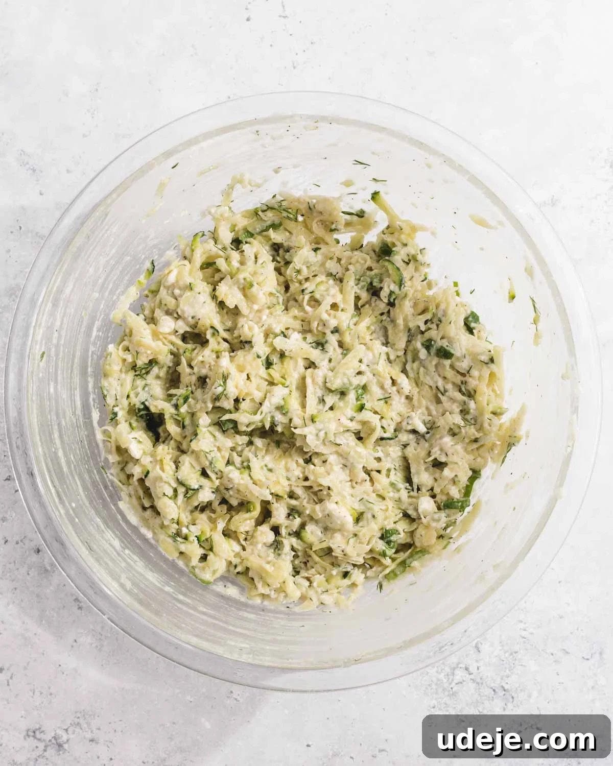 Crispy Turkish Zucchini Feta Fritters 7 A clear glass bowl filled with a mixture of shredded zucchini, herbs, and cheese sits on a light, textured surface. The mixture appears moist and well combined, ready for cooking or baking.