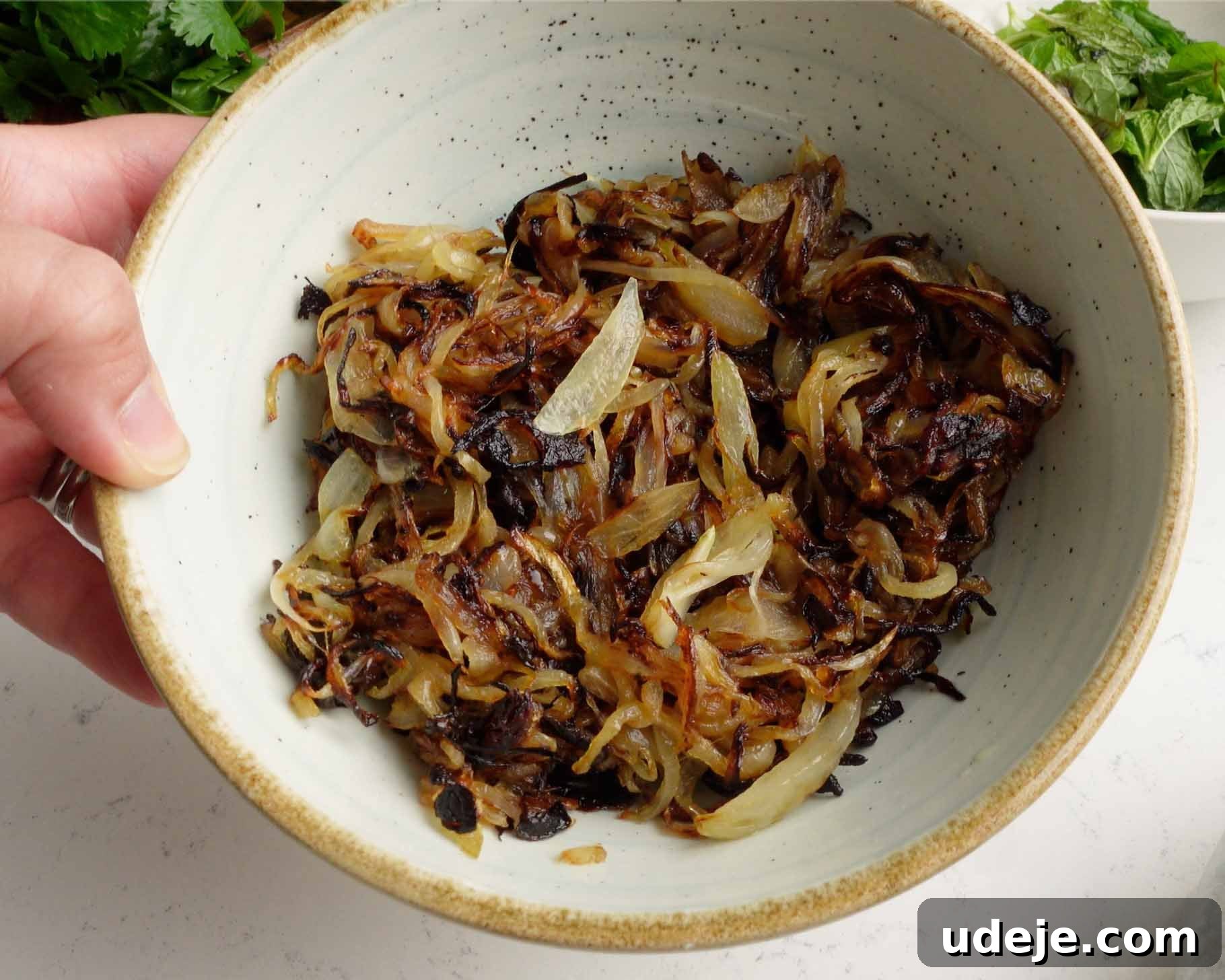 Melt-in-Your-Mouth Yemeni Lamb Mandi 11 A hand holds a speckled ceramic bowl filled with caramelized onions, golden brown and slightly charred, on a light countertop with greens visible in the background.