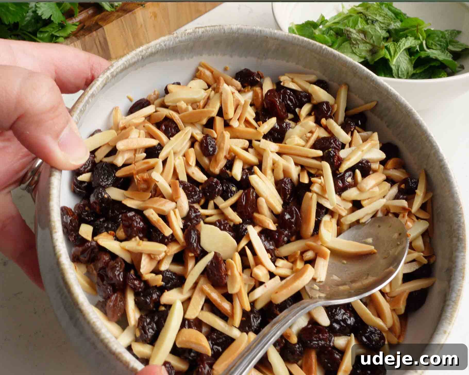 Melt-in-Your-Mouth Yemeni Lamb Mandi 12 A hand holds a bowl filled with slivered almonds and raisins, with a metal spoon inside. In the background, there is a white bowl containing fresh green herbs or lettuce leaves.