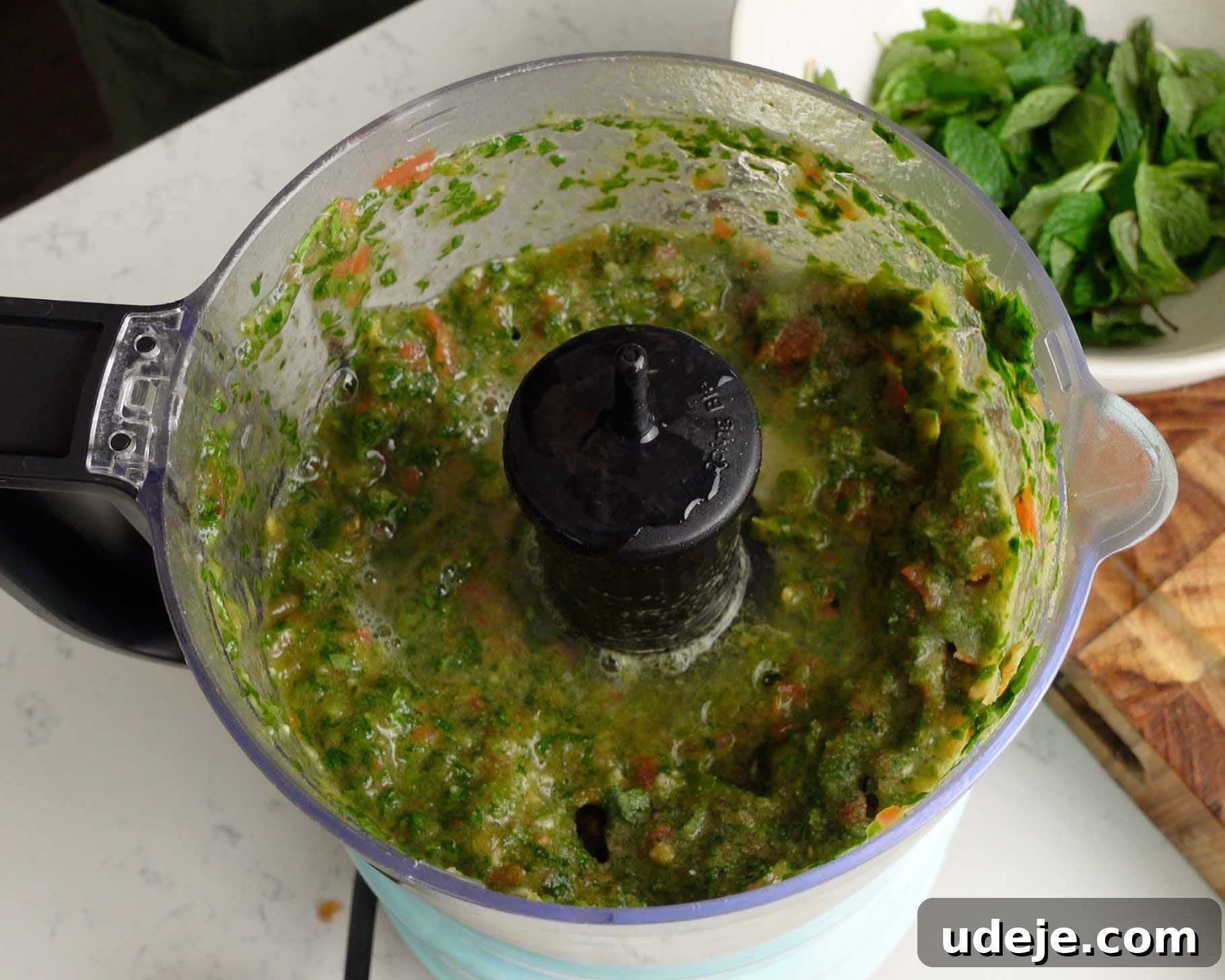 Melt-in-Your-Mouth Yemeni Lamb Mandi 13 A food processor containing a green, chunky mixture sits on a counter. Fresh mint leaves and a wooden cutting board are visible in the background.