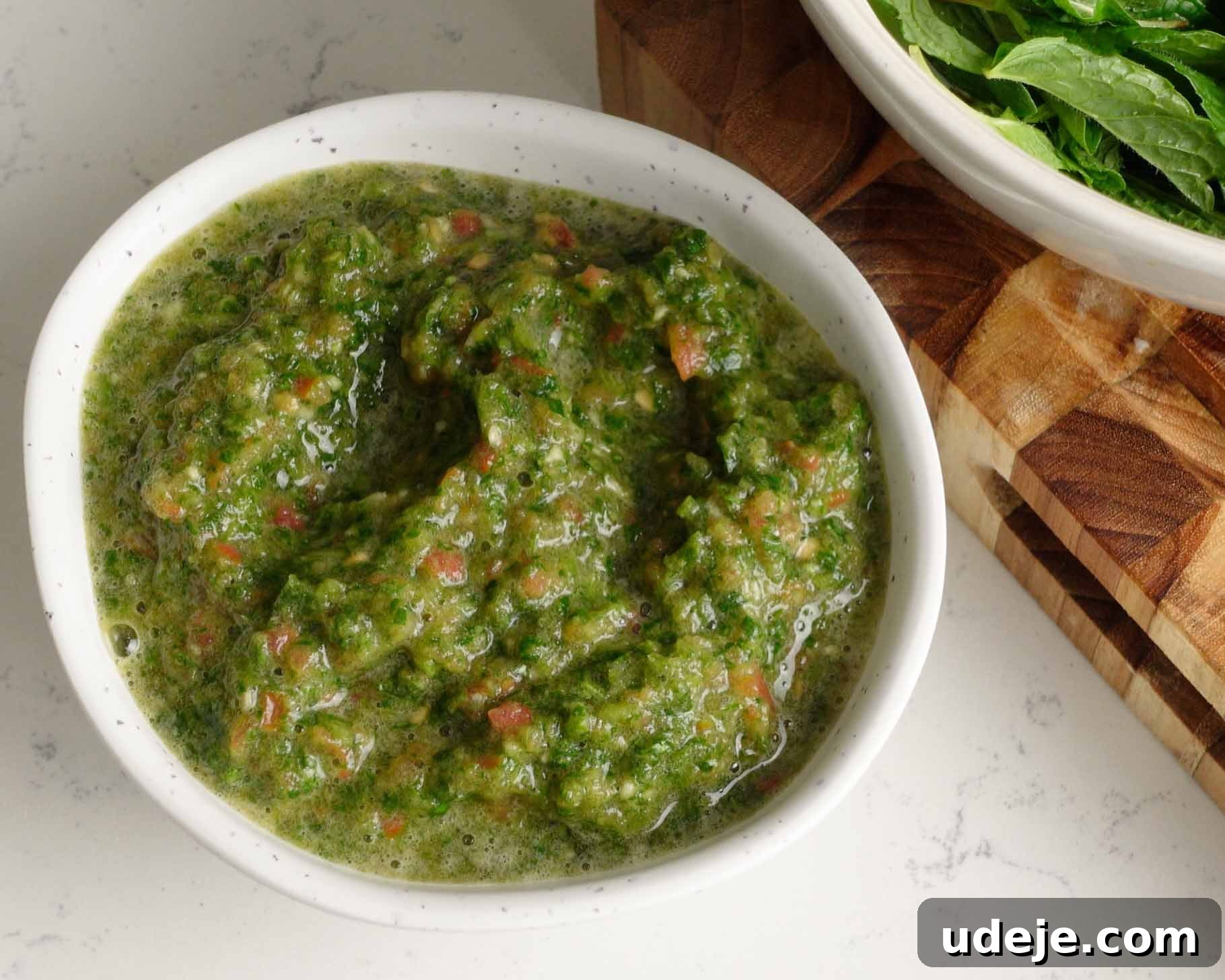 Melt-in-Your-Mouth Yemeni Lamb Mandi 14 A white bowl filled with chunky green sauce, placed on a light countertop next to a wooden cutting board and a bowl of leafy greens.