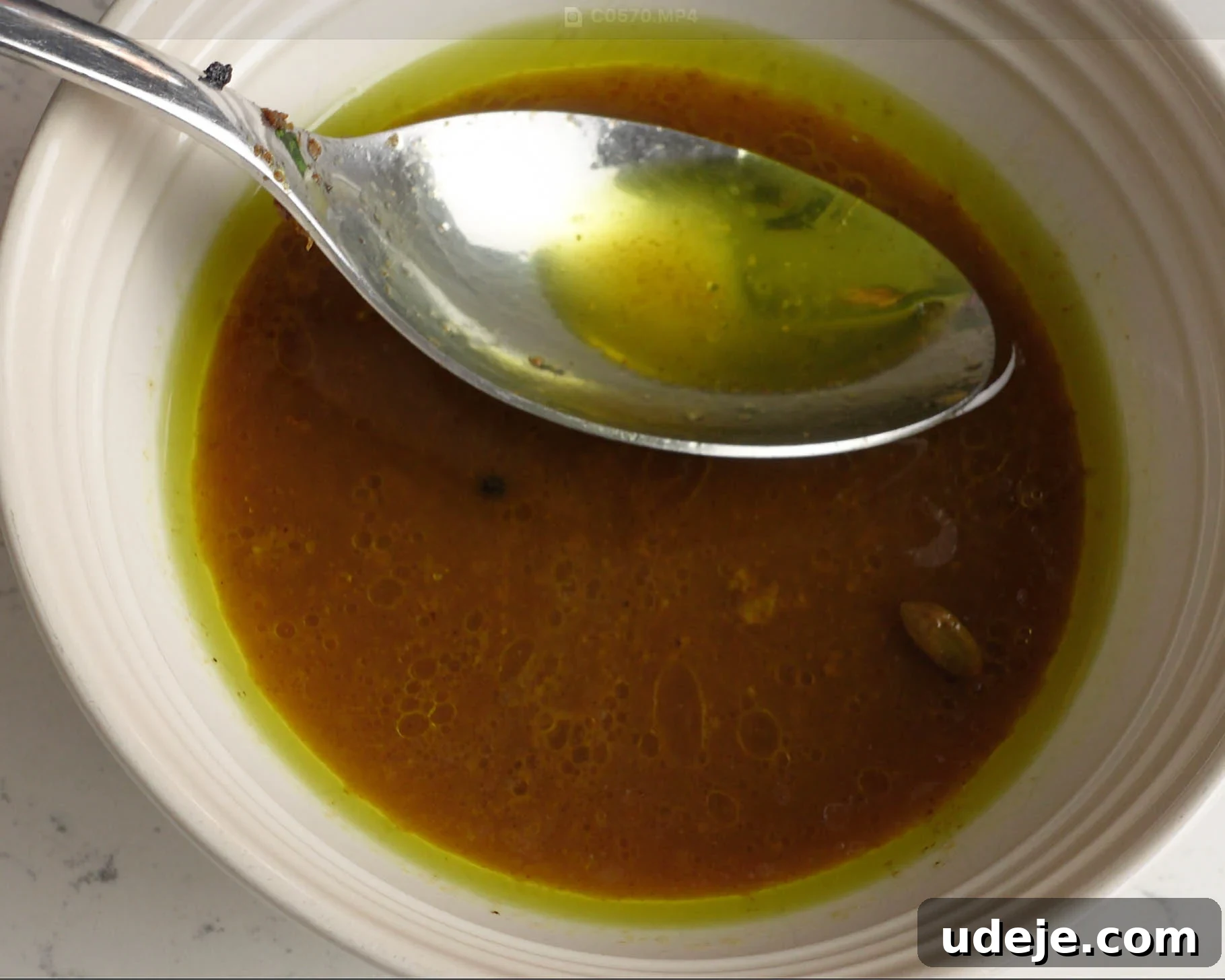 Melt-in-Your-Mouth Yemeni Lamb Mandi 15 A close-up of a white bowl containing an oily, golden-brown liquid with visible spices. A metal spoon rests inside, reflecting the green-tinted oil on its surface.